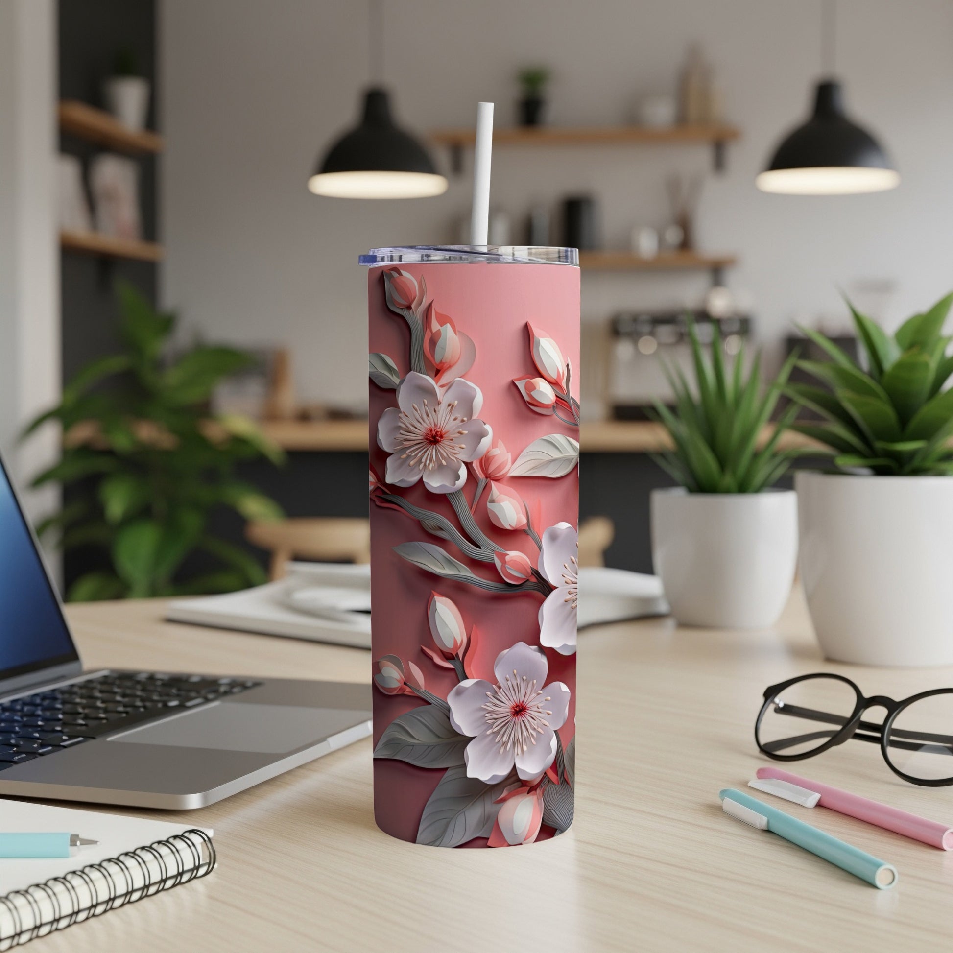 Pink floral tumbler with straw on a desk, next to a laptop and pens.