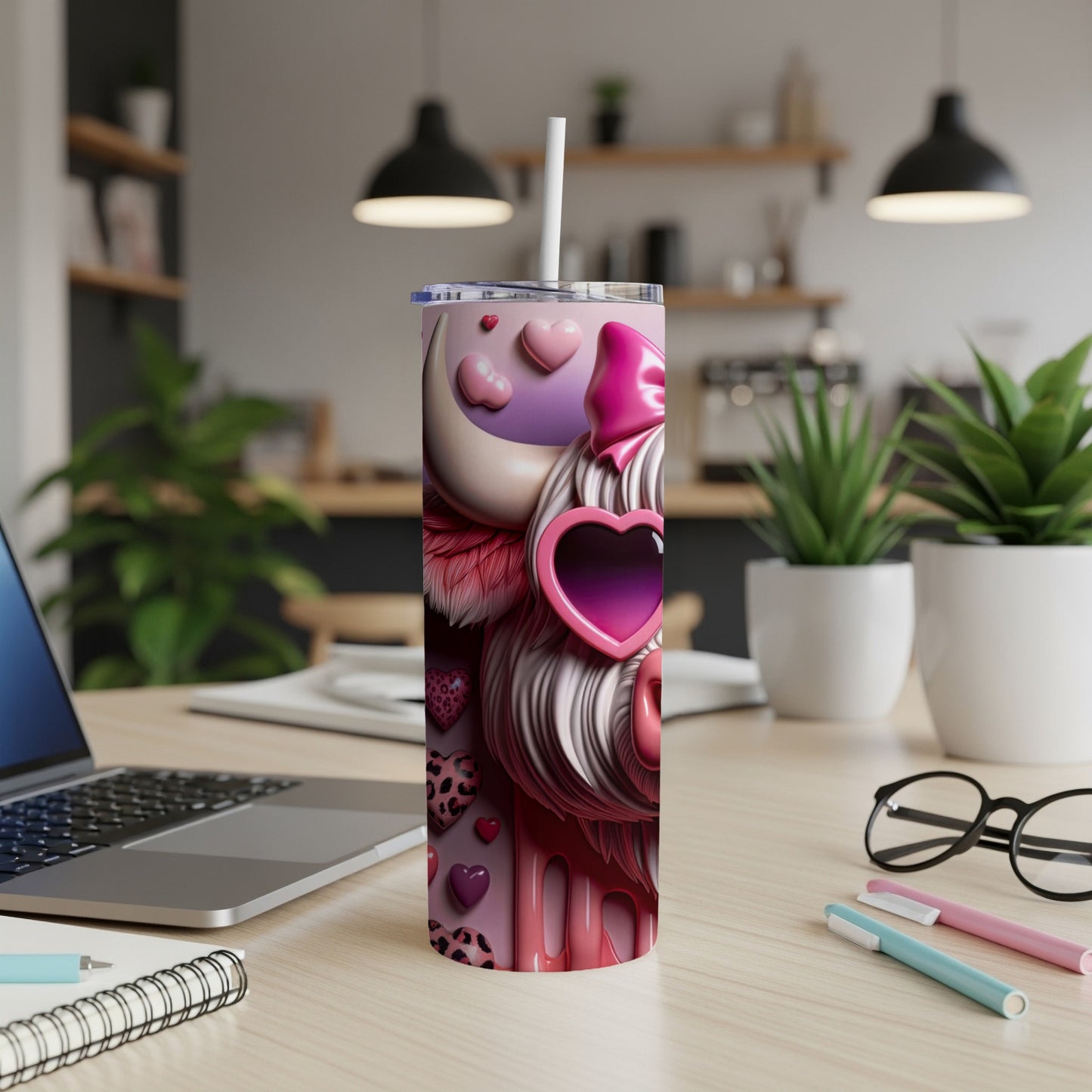 Pink tumbler with hearts and a bow on a desk with a laptop and pens.
