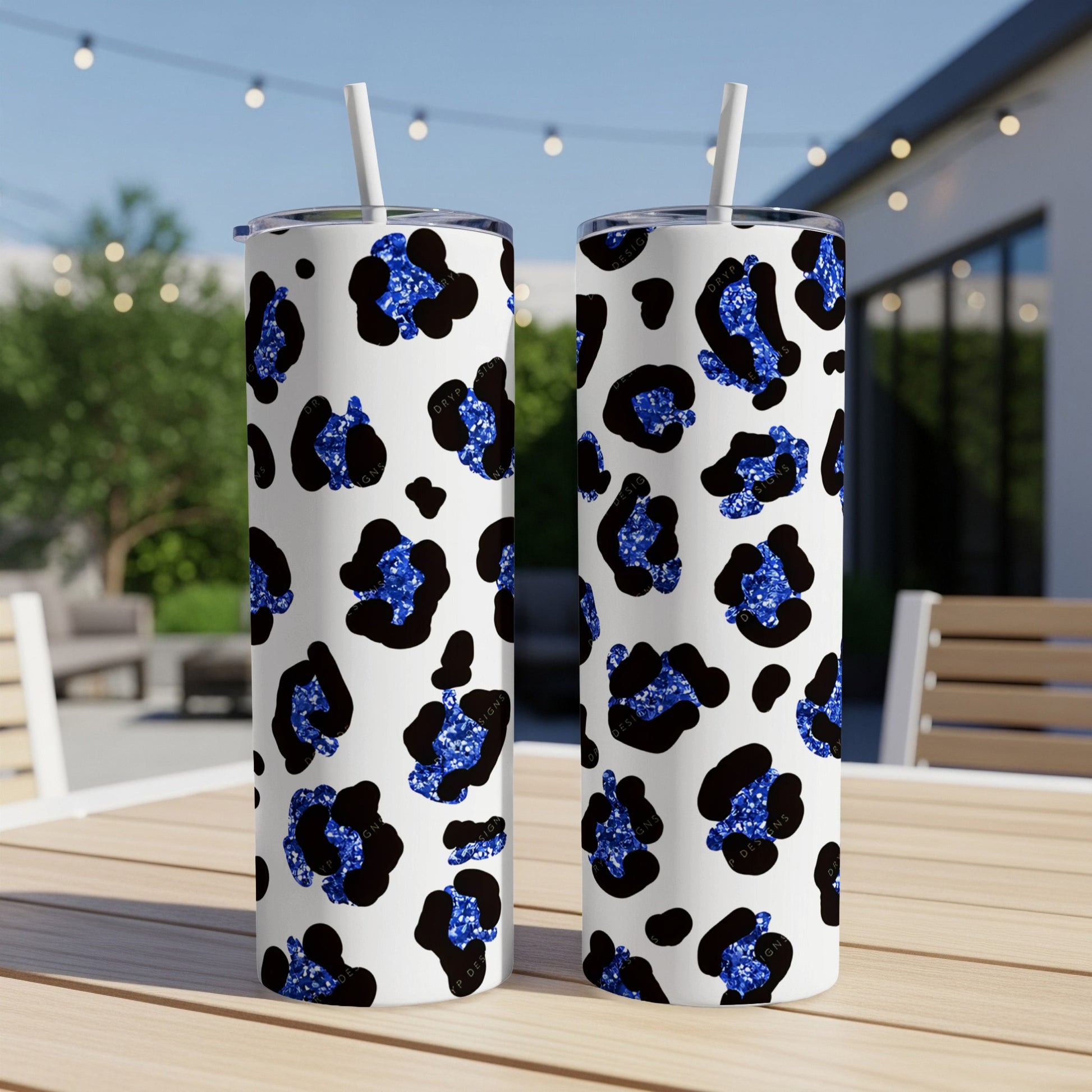 Two white tumblers with blue glitter leopard print, on a table outdoors.