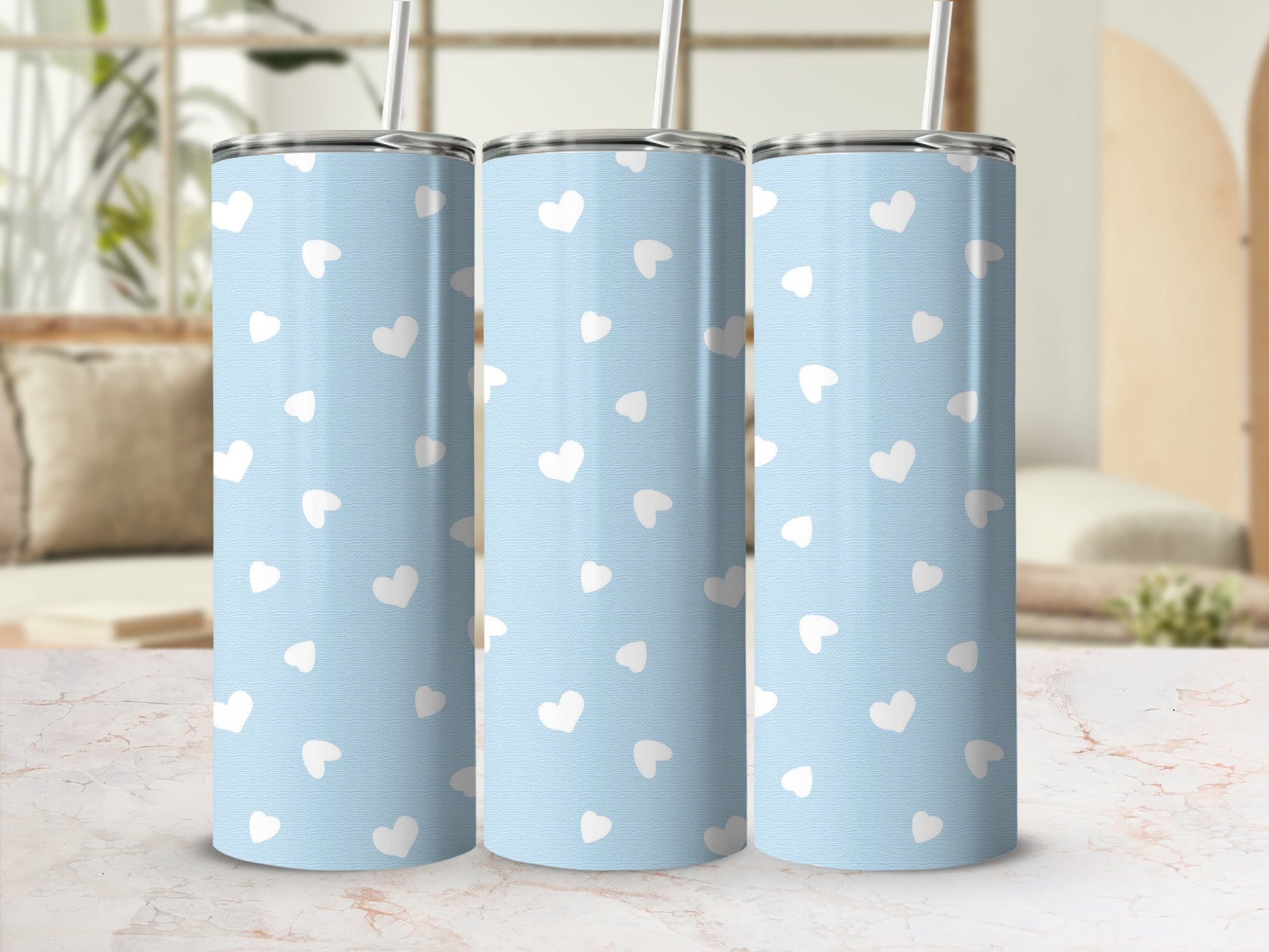 Three blue tumblers with white heart pattern and silver lids with straws on marble counter.