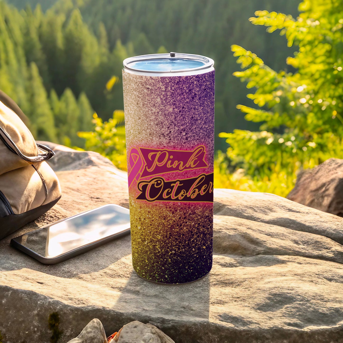 Pink October tumbler with glitter gradient, resting on rocks with backpack and phone.