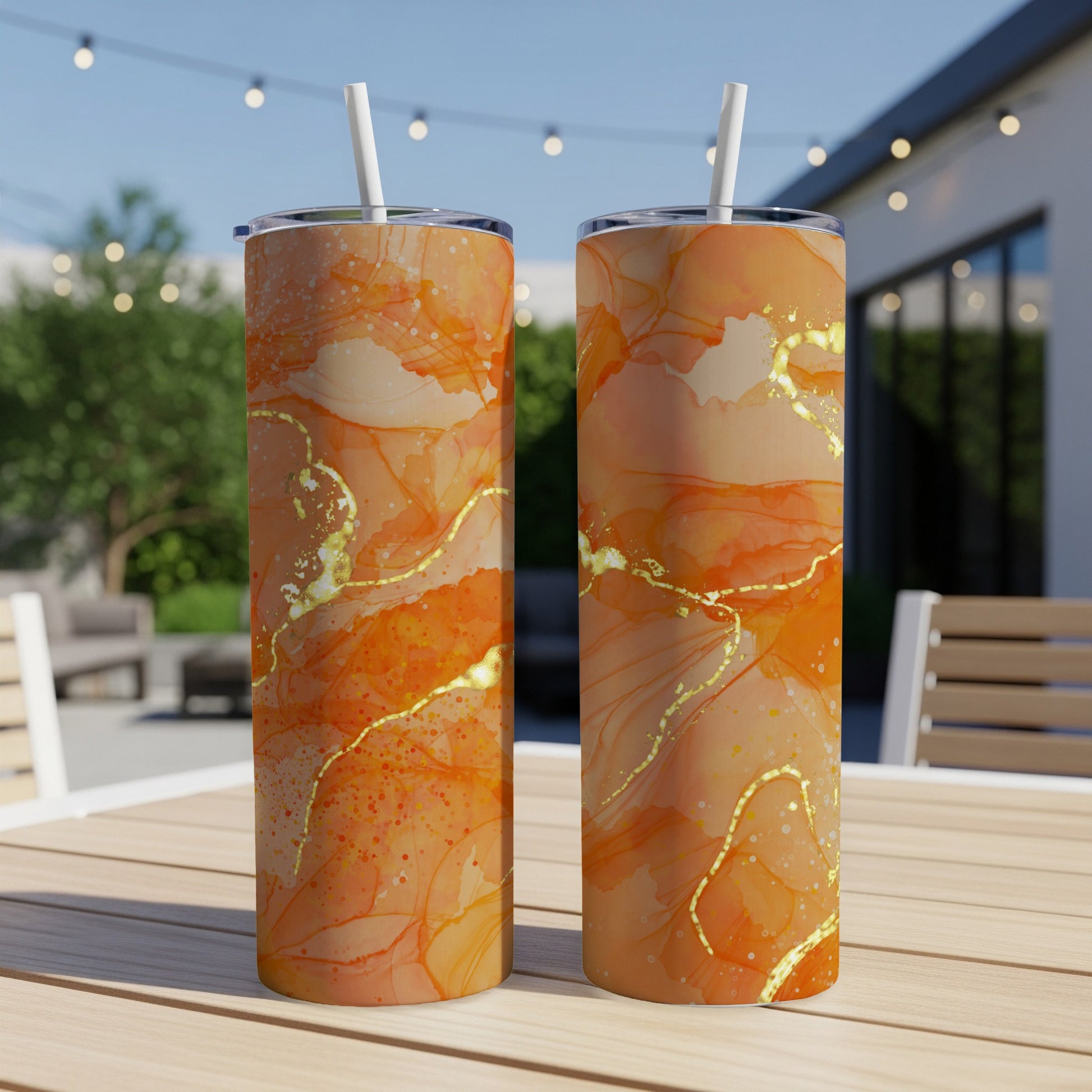 Two orange and gold marbled tumblers with straws on a wooden table outdoors.