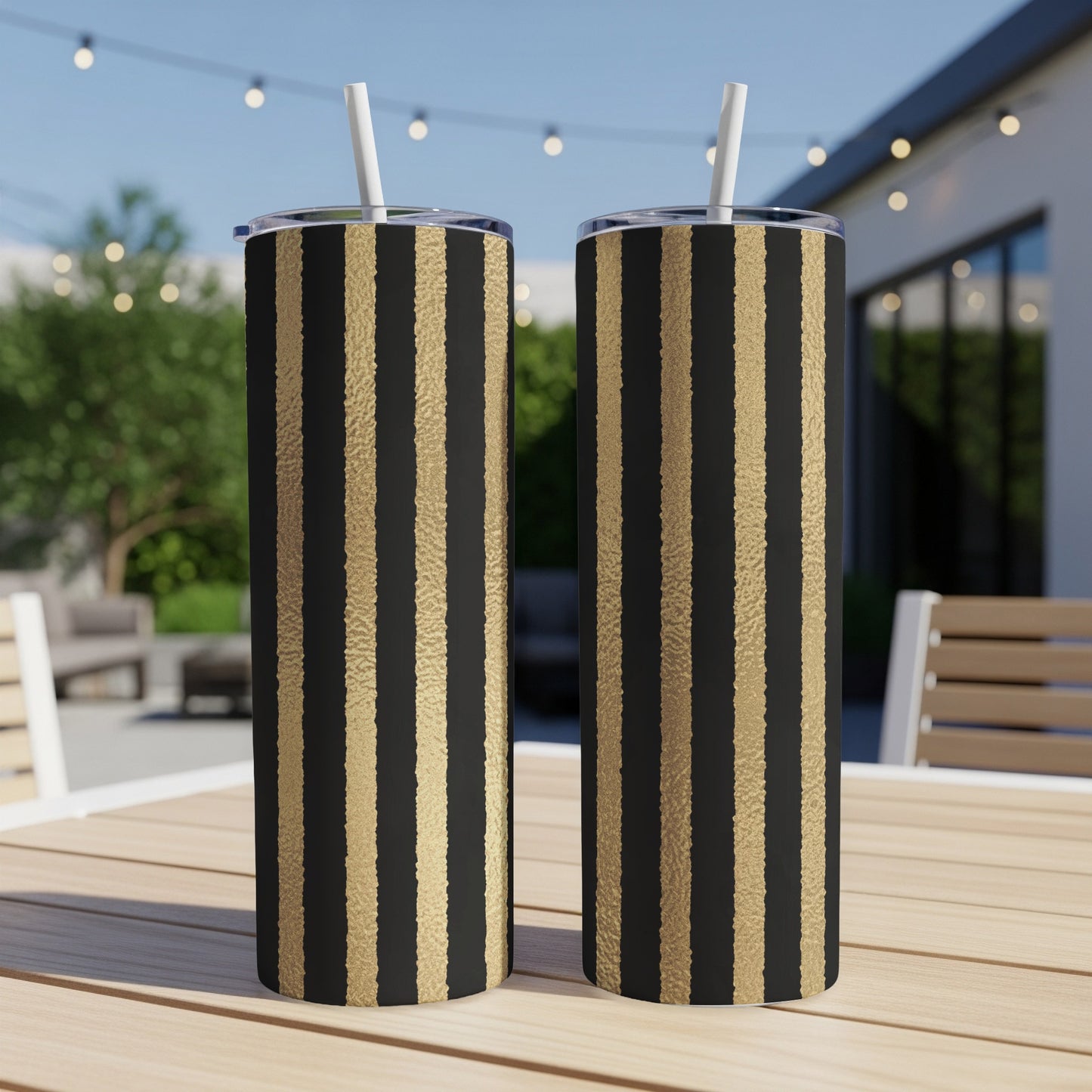 Two tumblers with black and gold vertical stripes, each with a straw, sit on a wooden table outdoors.