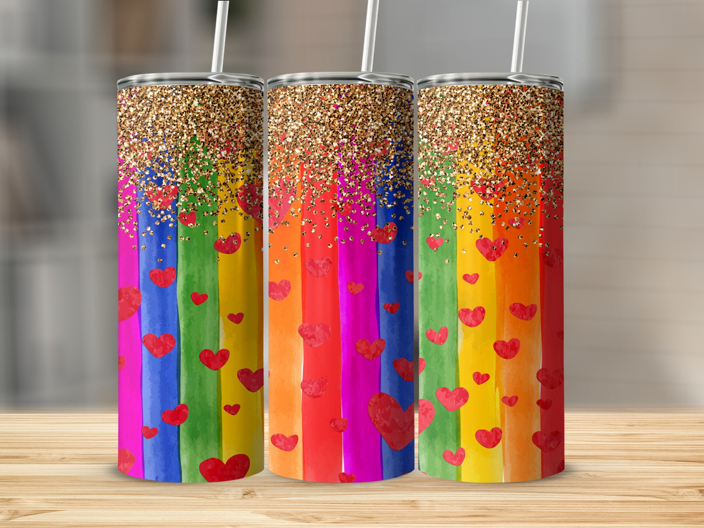 Three rainbow tumblers with glitter and heart designs on a wooden surface.