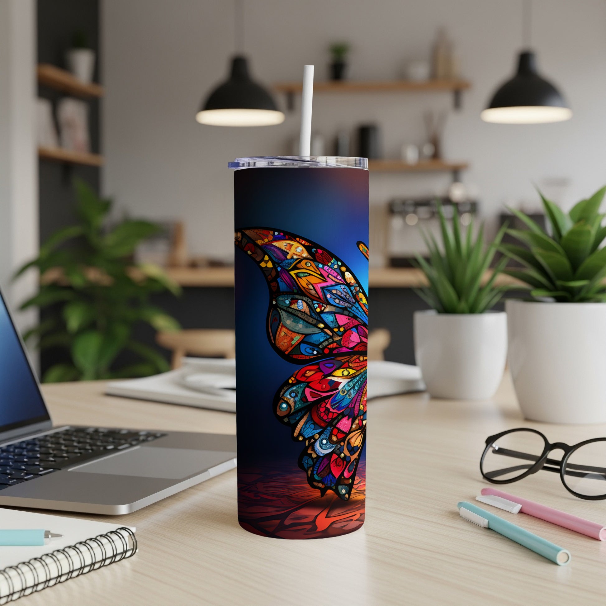 Colorful butterfly tumbler with straw on desk beside laptop and pens.
