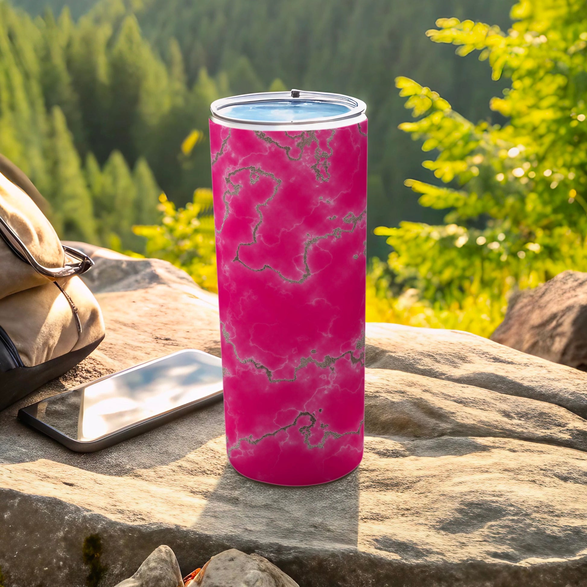 Pink marble tumbler with silver accents on a rock, beside a backpack and phone.