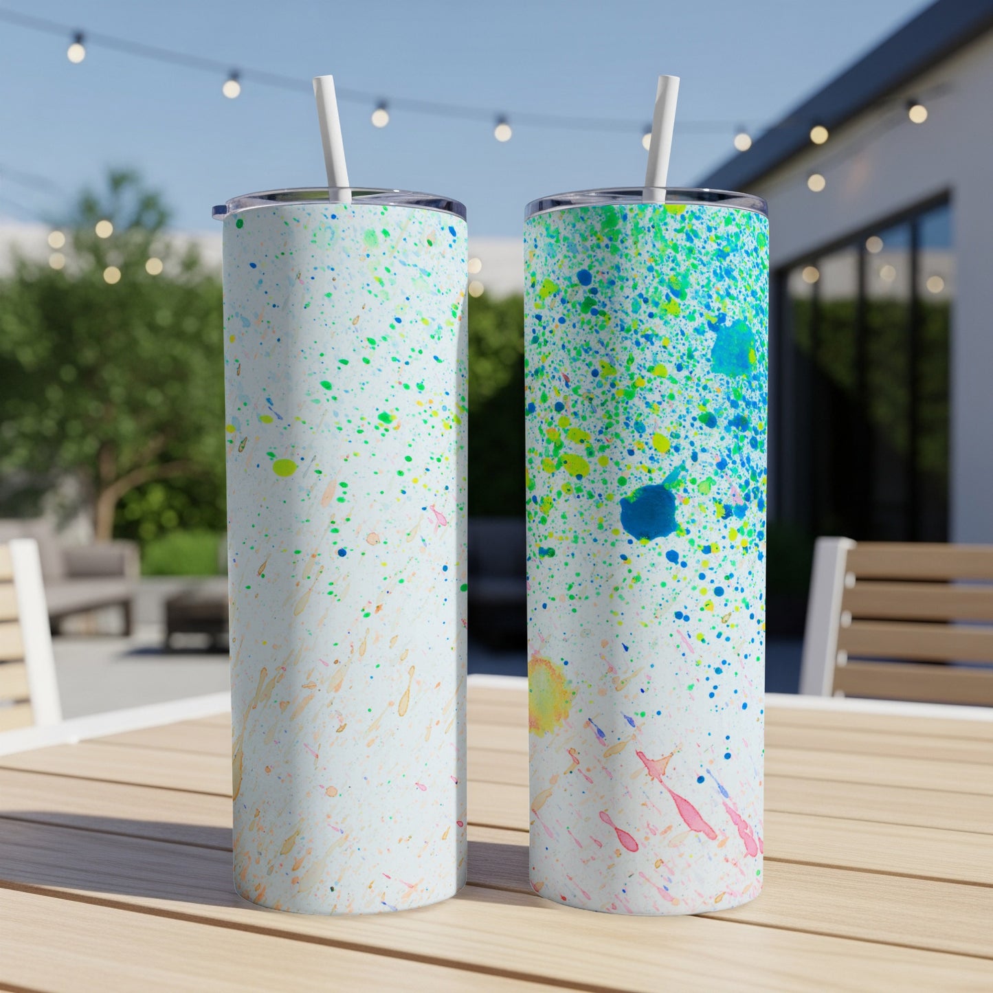 Two tumblers with colorful paint splatters on a wooden table outdoors.