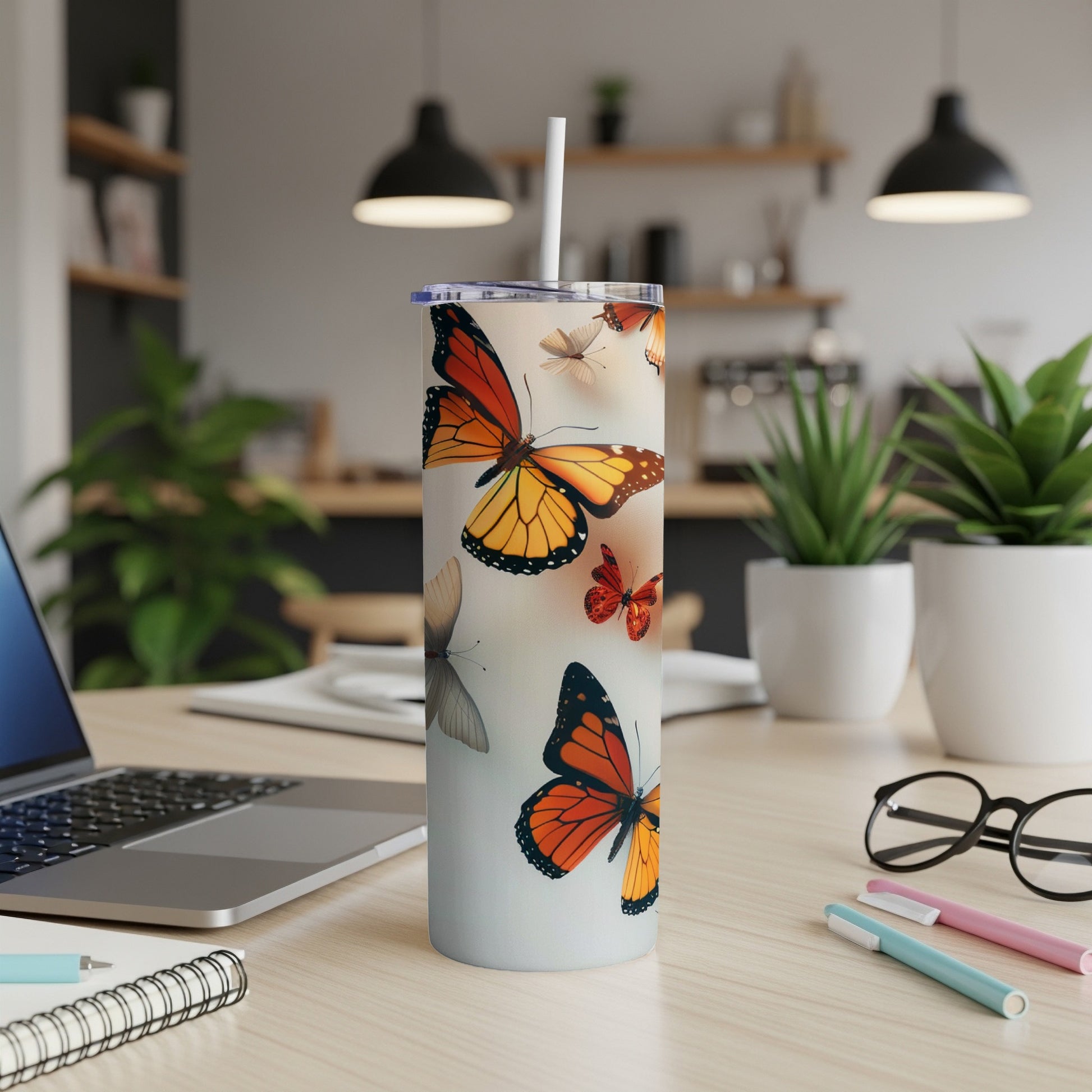 Tall tumbler with monarch butterflies, straw, laptop, and pens on a desk.