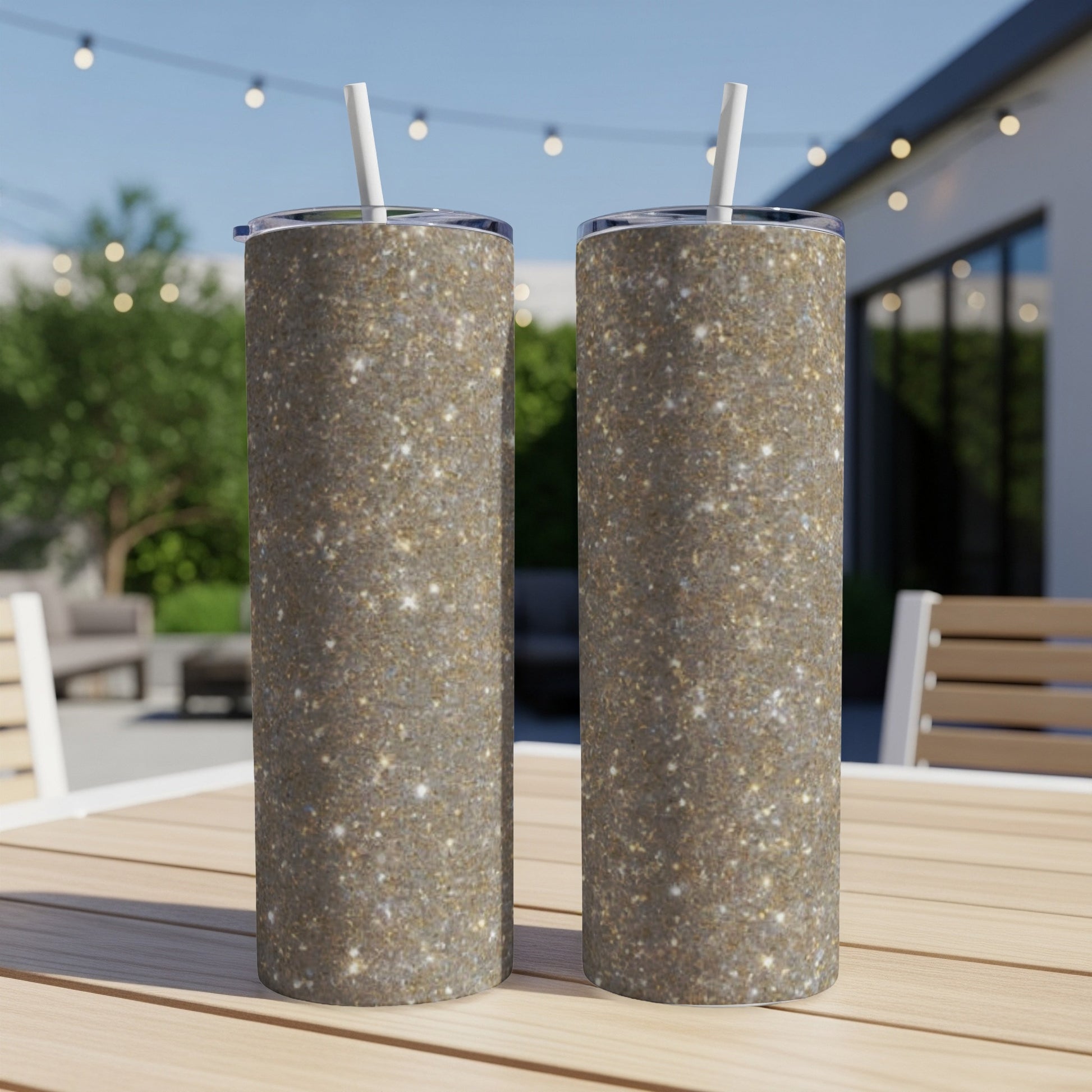 Two glitter-covered tumblers with straws sit on a wooden table outdoors.