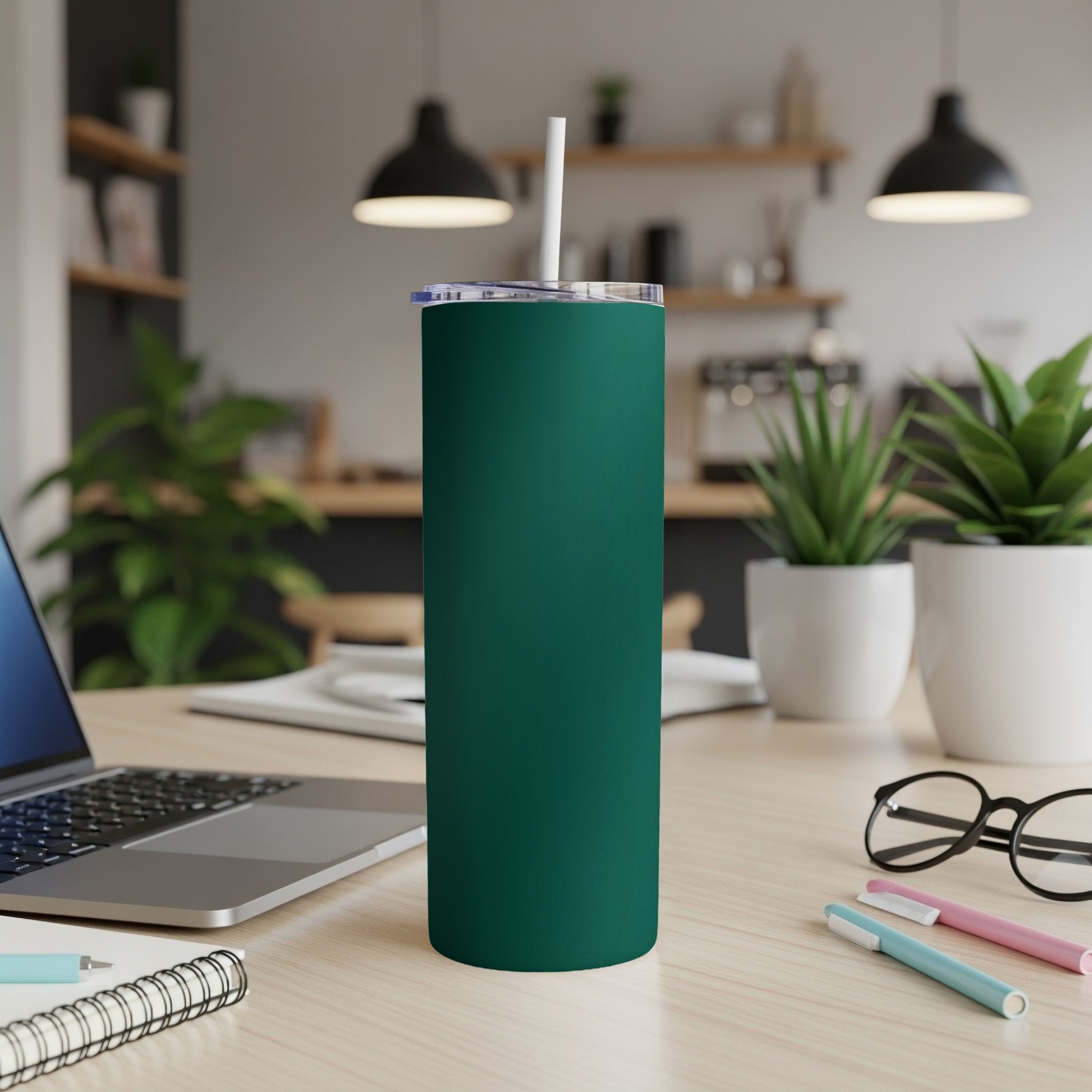 Dark teal tumbler with white straw on a wooden desk with laptop and plants.