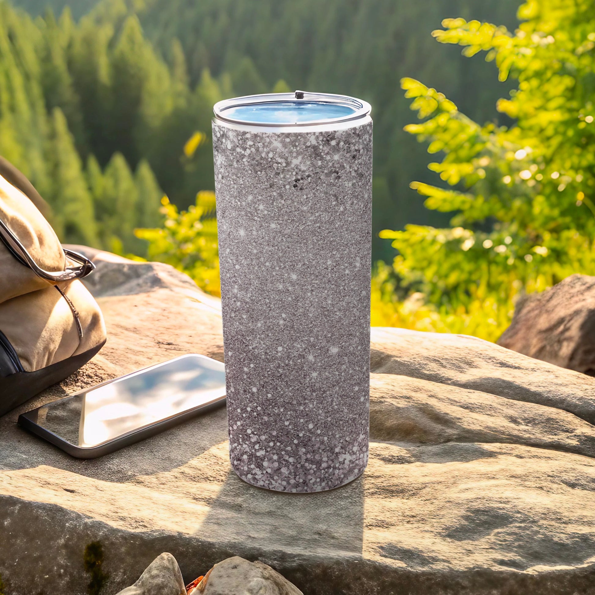 Sparkling silver glitter tumbler on rock with phone and backpack.