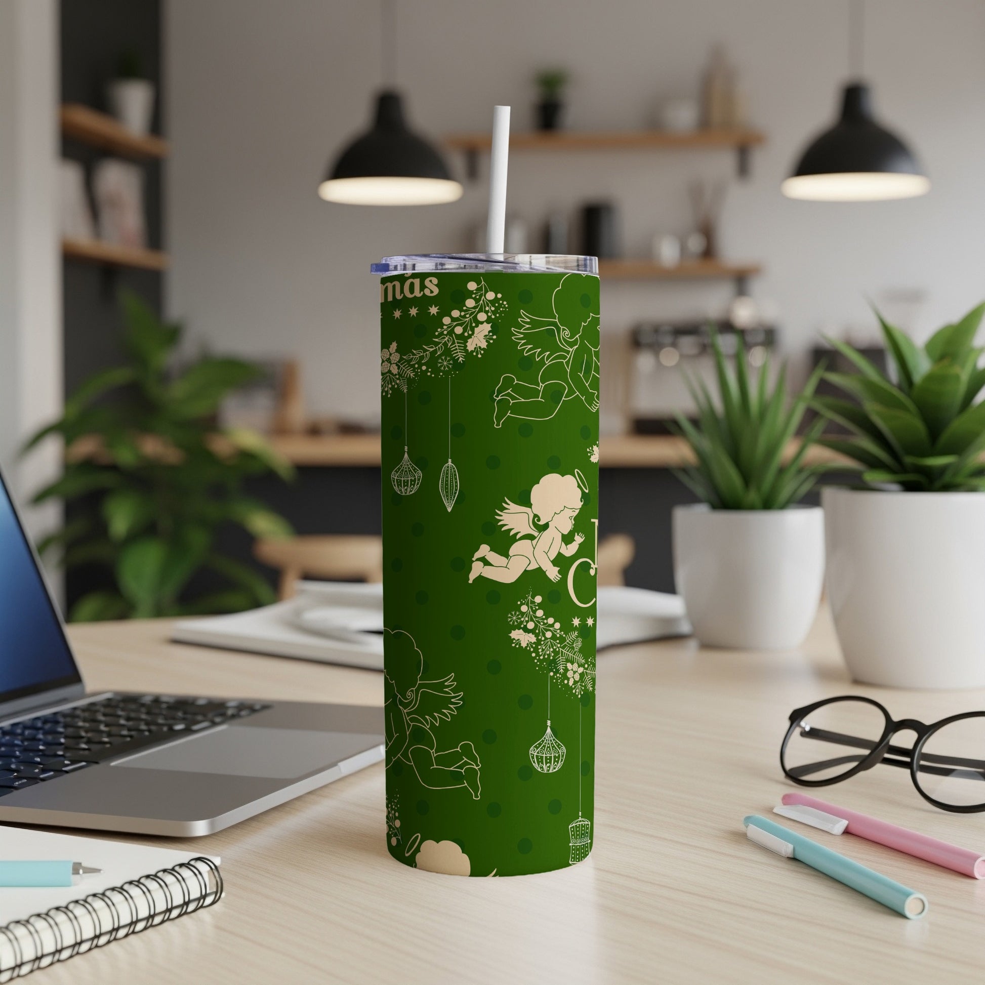 Green tumbler with white Christmas angels and ornaments, on a desk.