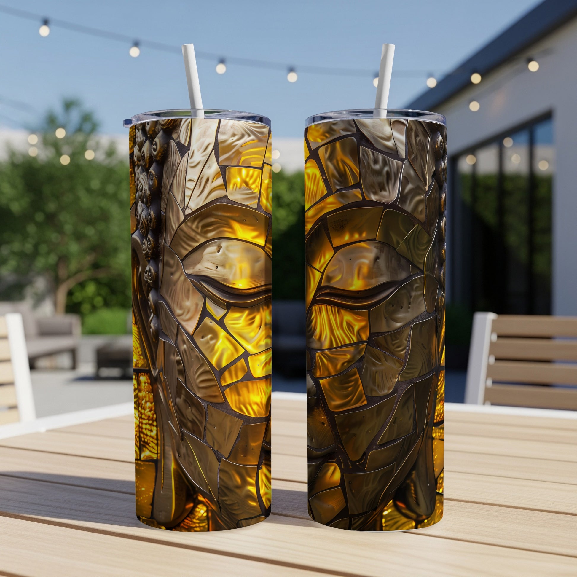 Two golden, mosaic-style Buddha face tumblers with straws on a wooden table.