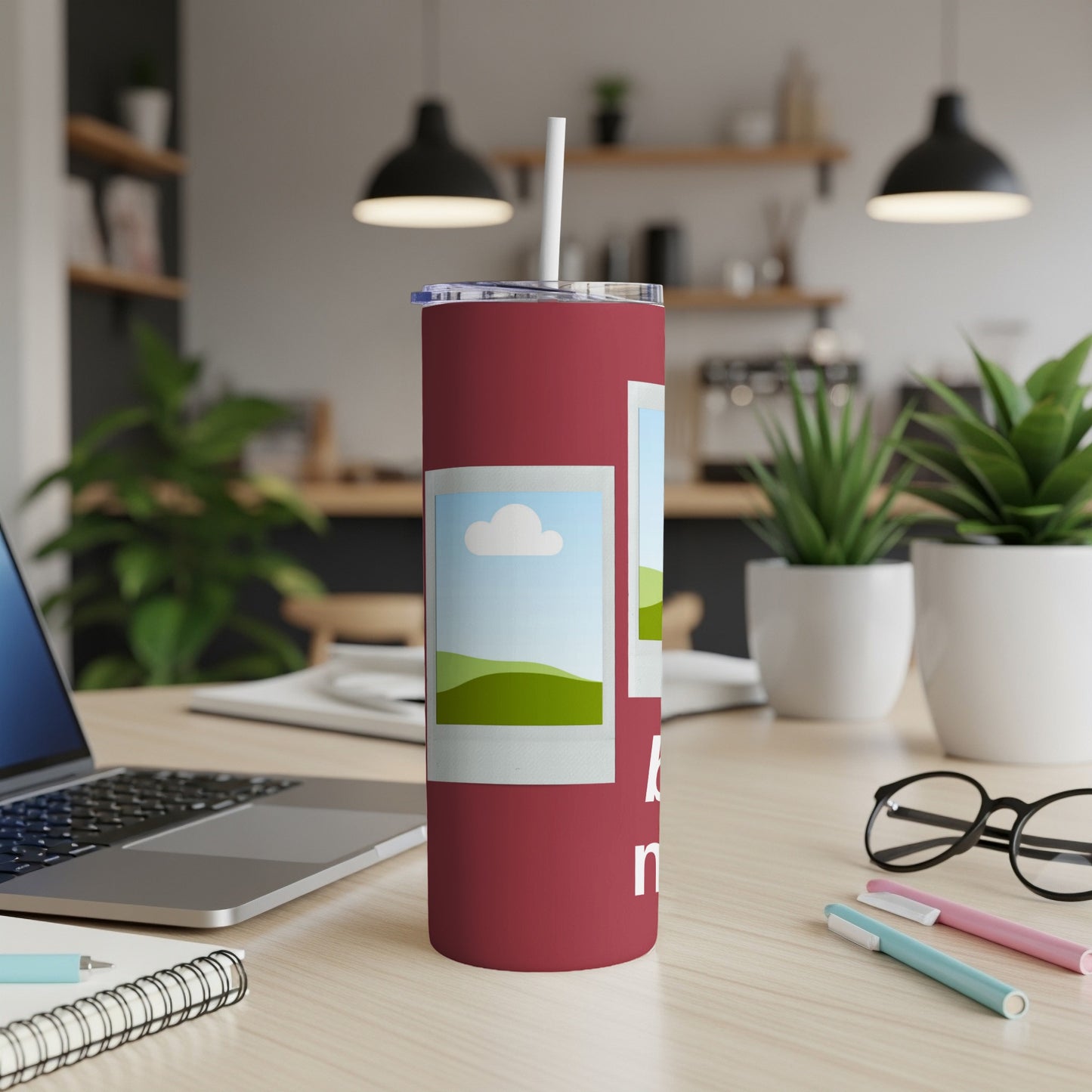 Maroon tumbler with two framed landscape images and partial text on a desk.