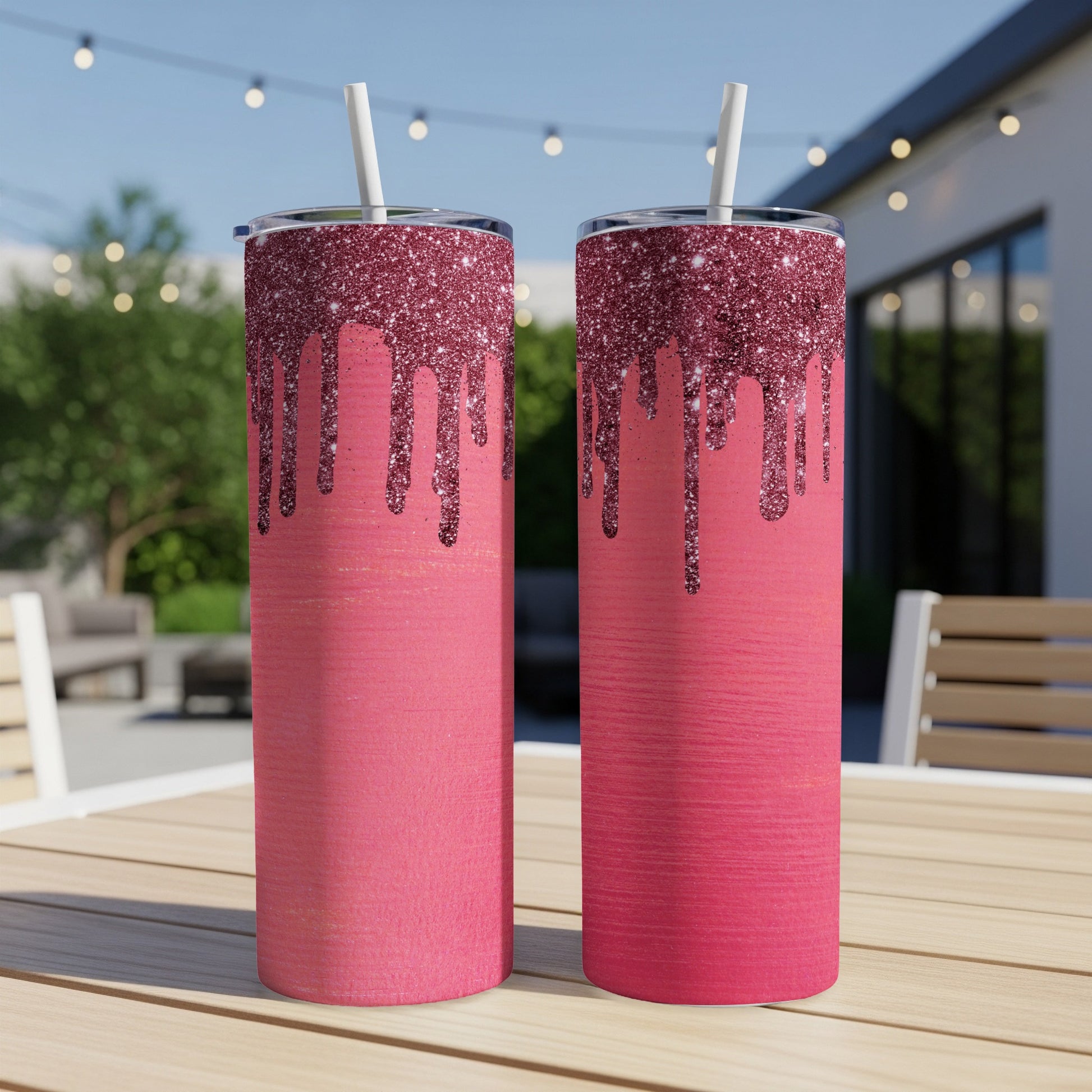 Two tall pink tumblers with glitter drip design sit on a wooden table outdoors.