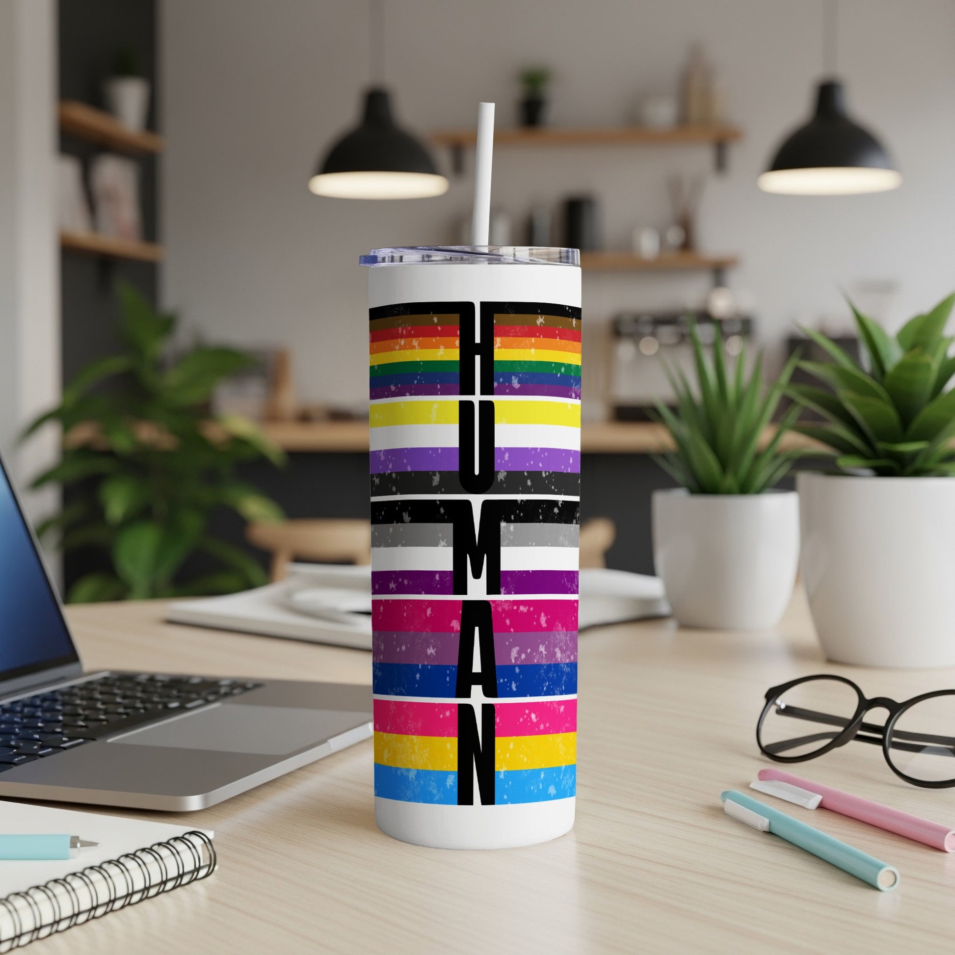 Pride flag tumbler with "HUMAN" text, laptop, and plants in background.