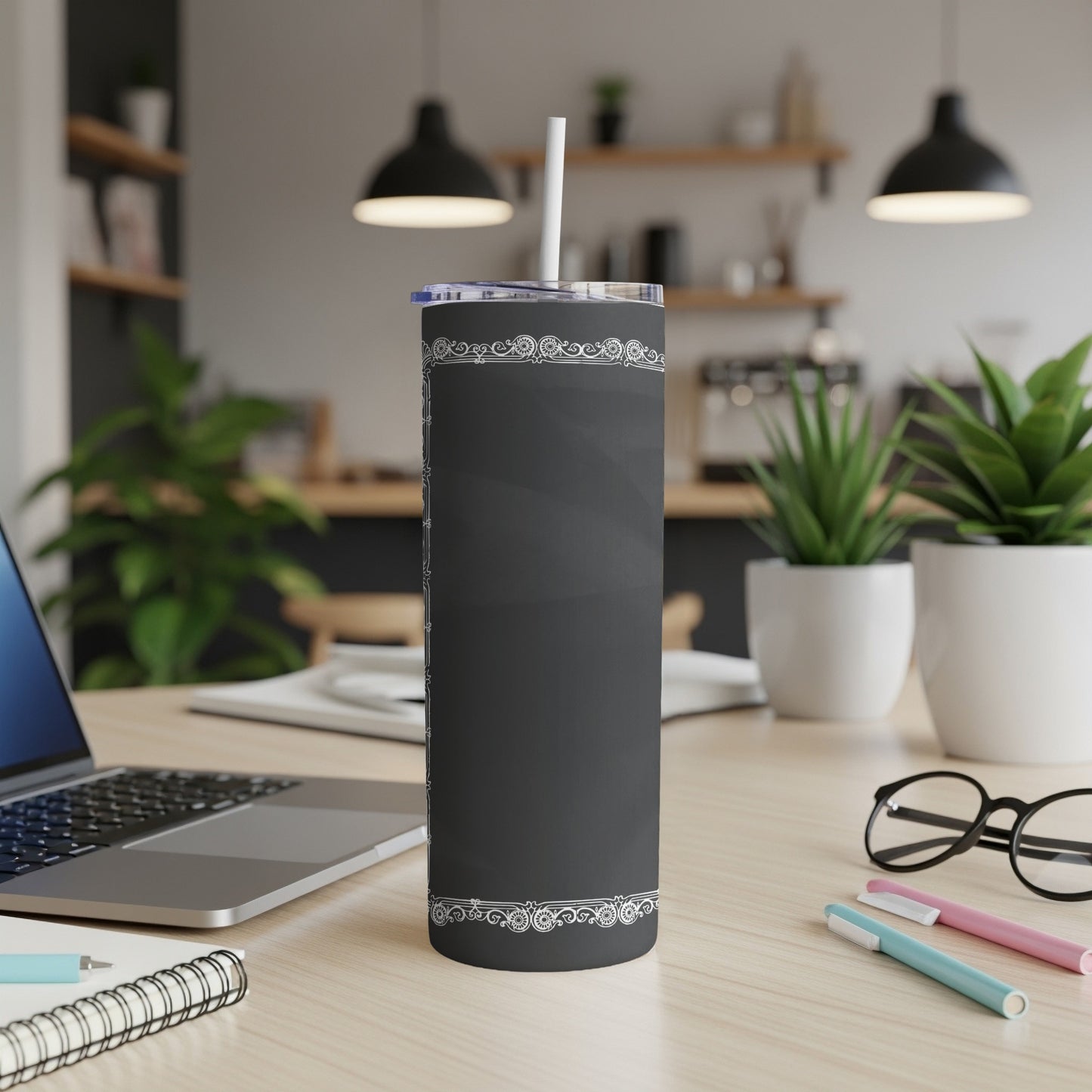 Black tumbler with white scrollwork design on a desk with a laptop and plants.