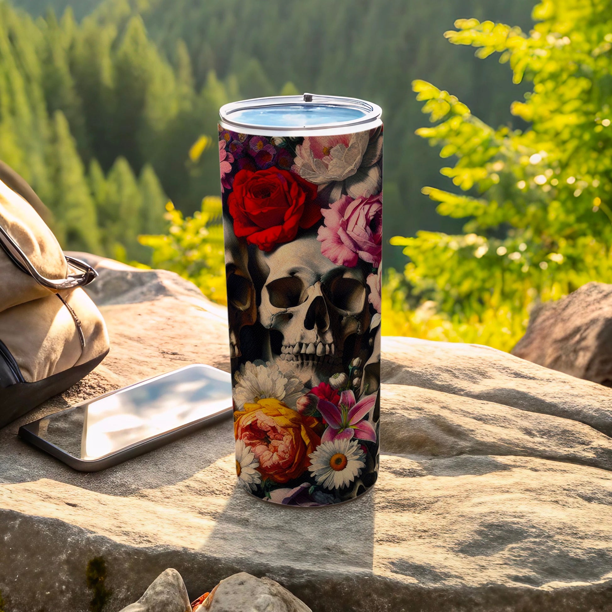 Tumbler with skulls and colorful flowers, resting on rocks outdoors.