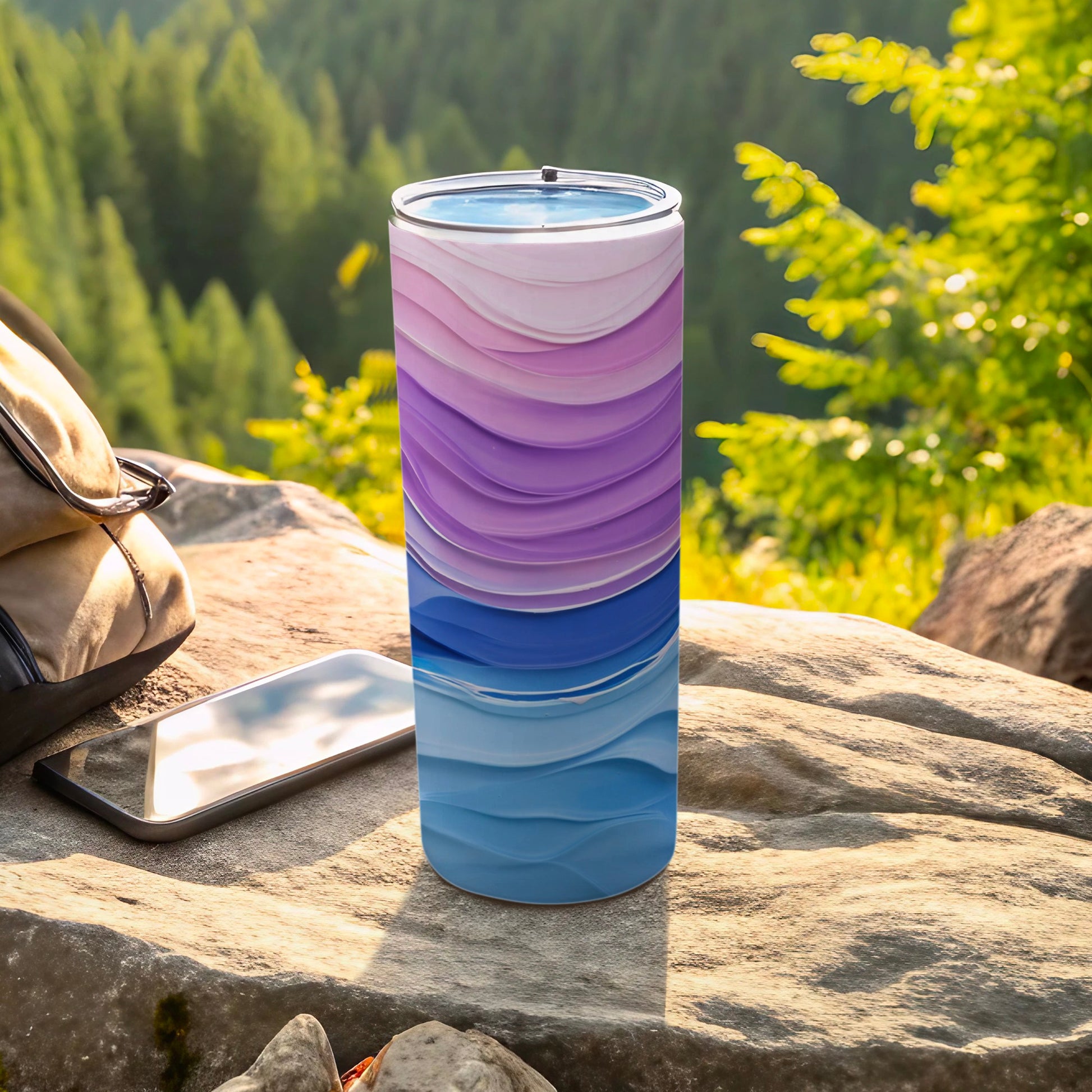 Tumbler with wave pattern of purple and blue layers on rock with phone and backpack.