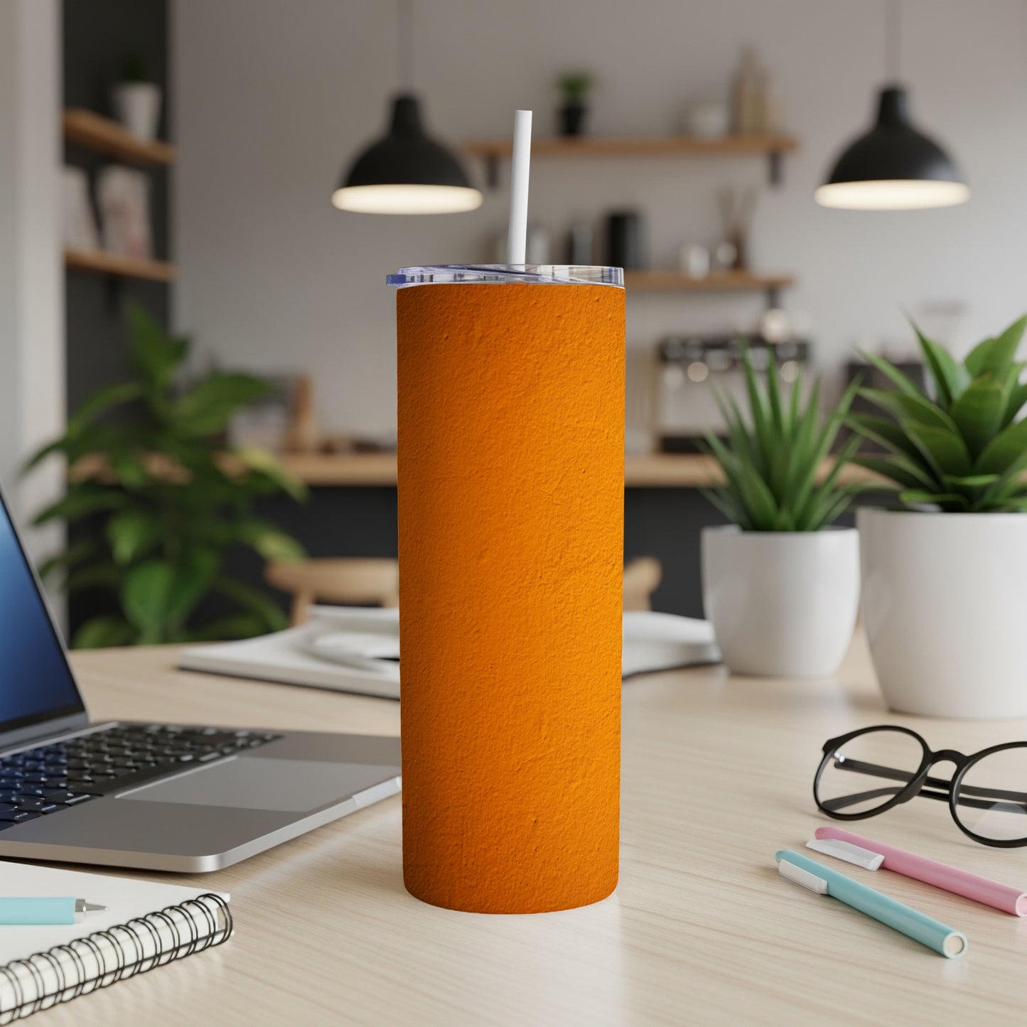 Orange textured tumbler with white straw on a desk with laptop and pens.