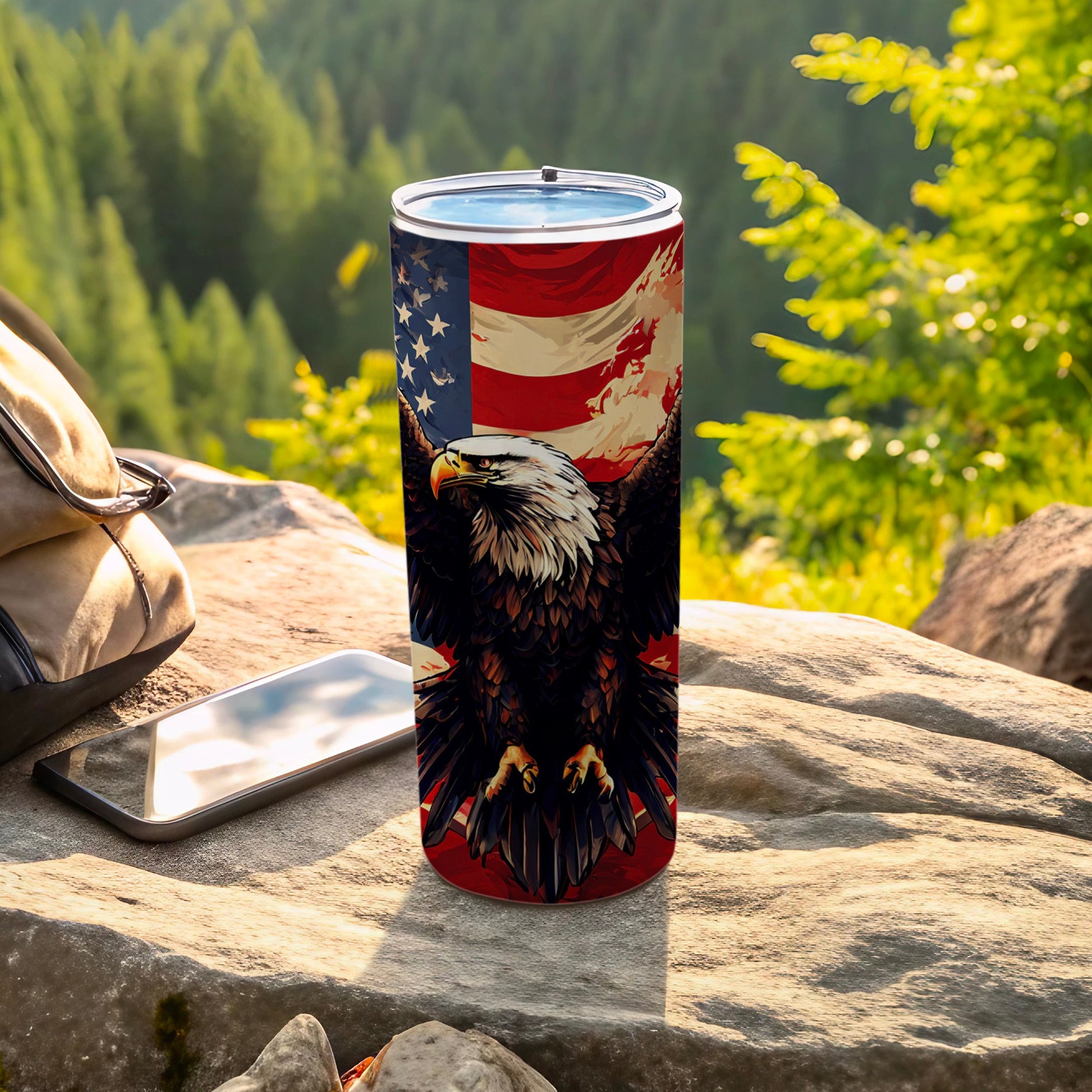 Eagle and US flag tumbler on rocky surface with backpack and phone.