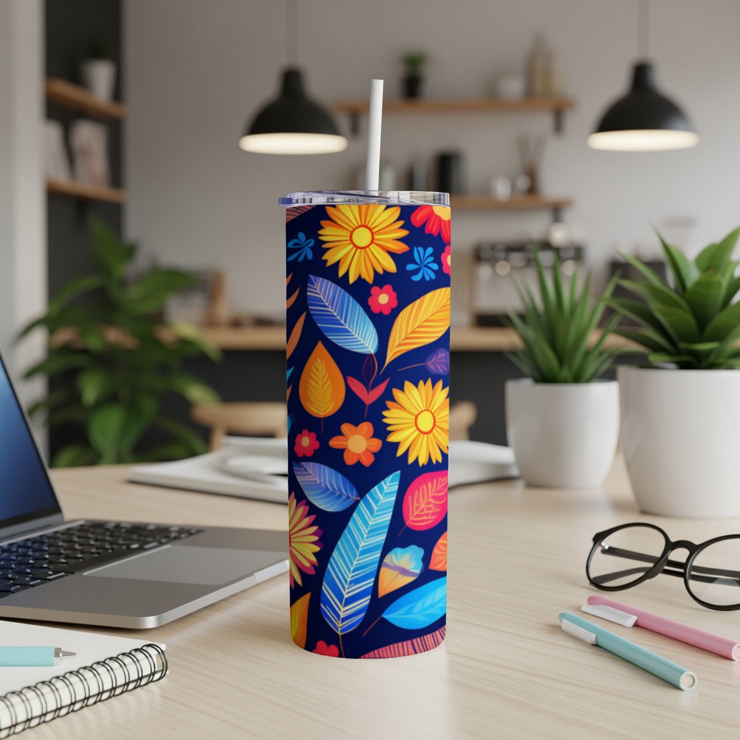 Blue tumbler with colorful floral and leaf pattern, with straw, on desk.