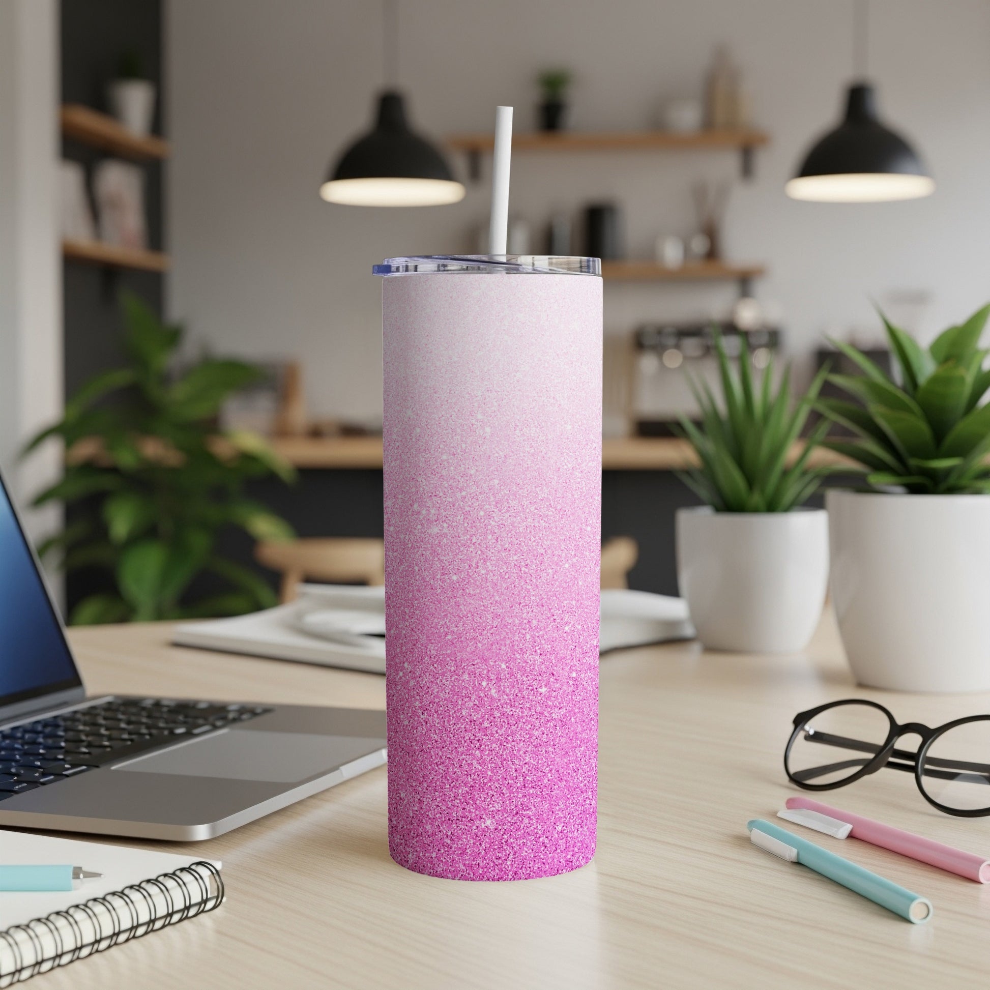 Pink glitter tumbler with straw on desk next to laptop and pens.