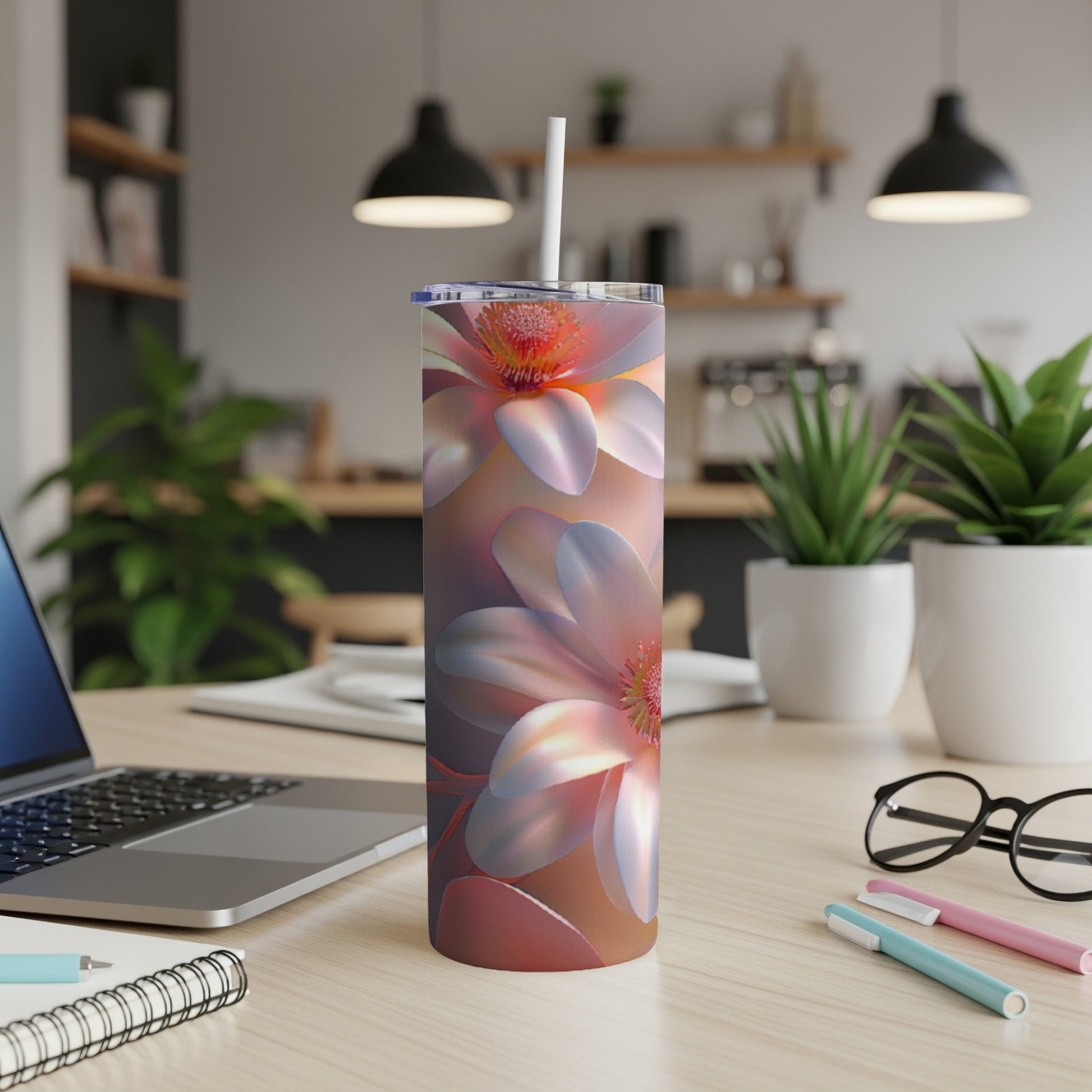 Floral tumbler with straw on a desk beside a laptop and pens.