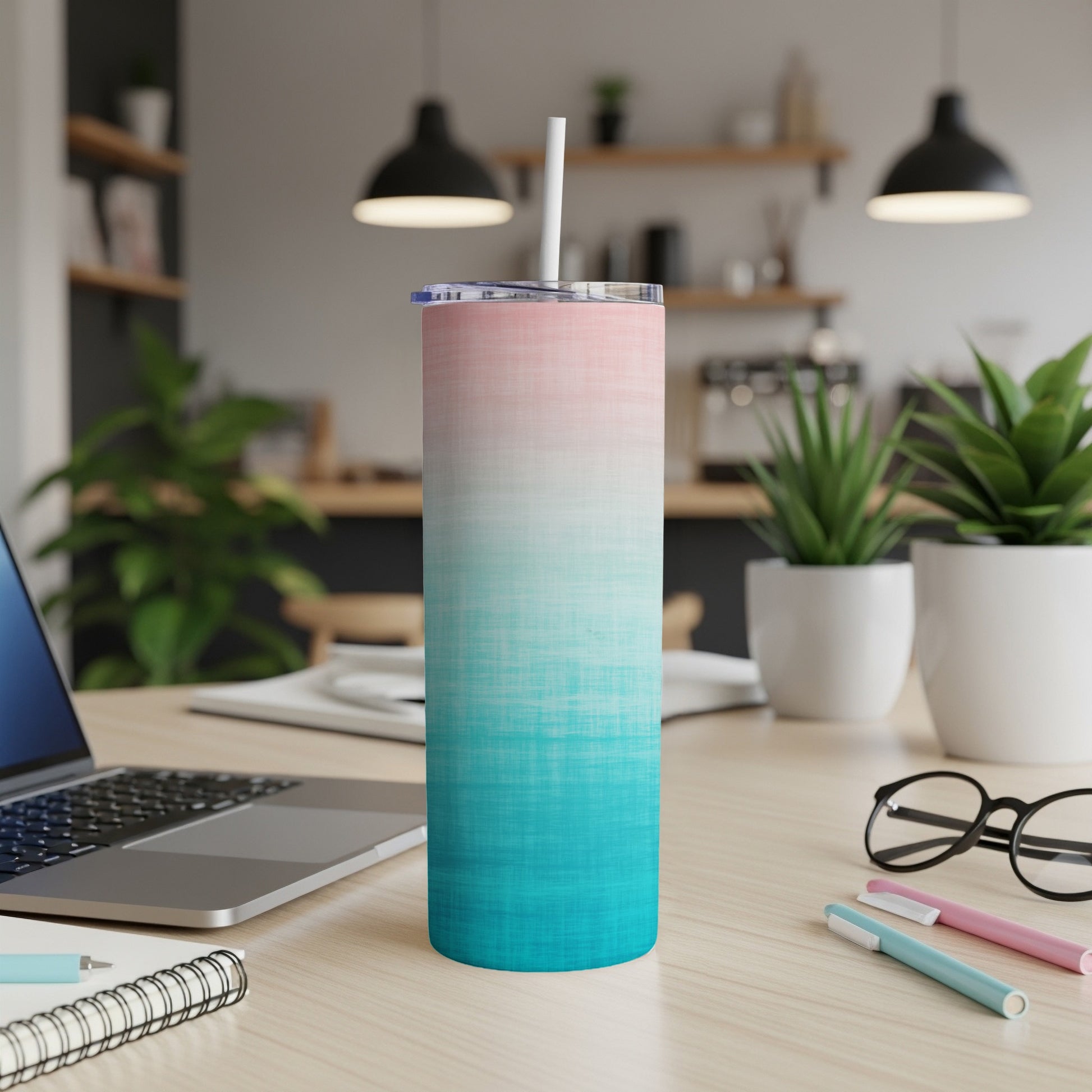 Gradient pink and teal tumbler with straw on desk next to laptop and pens.