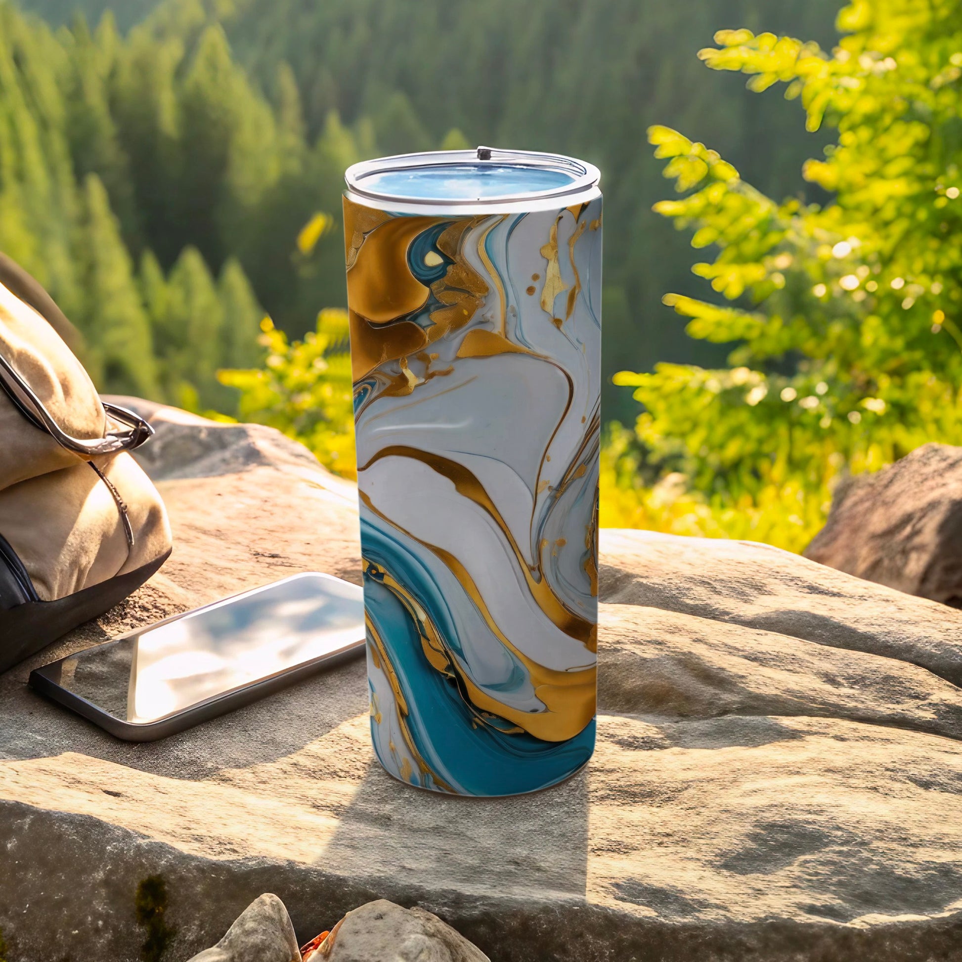 Tall tumbler with blue, gold, and white marble pattern on rock with phone and backpack.