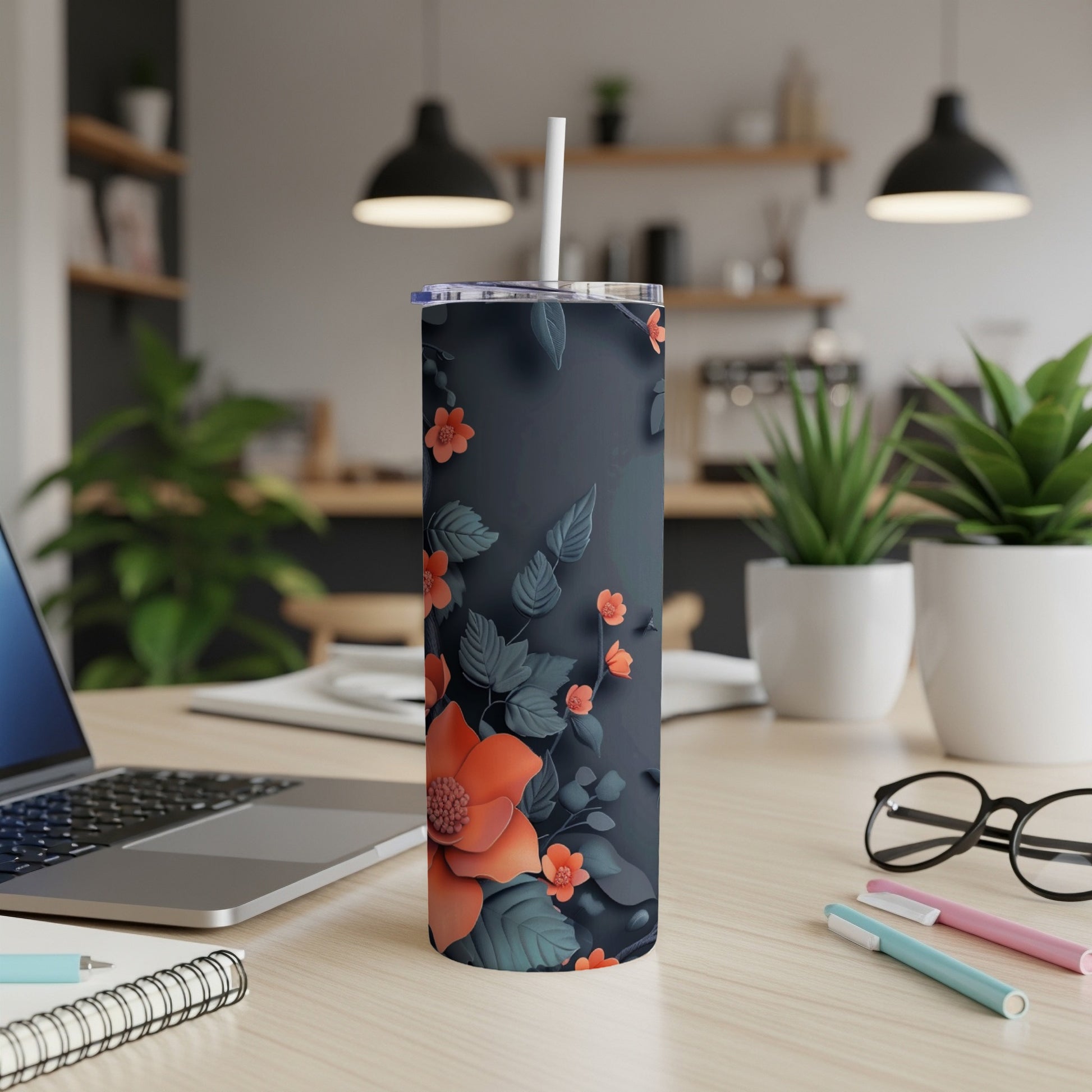 Dark tumbler with orange floral pattern on a desk with laptop and pens.