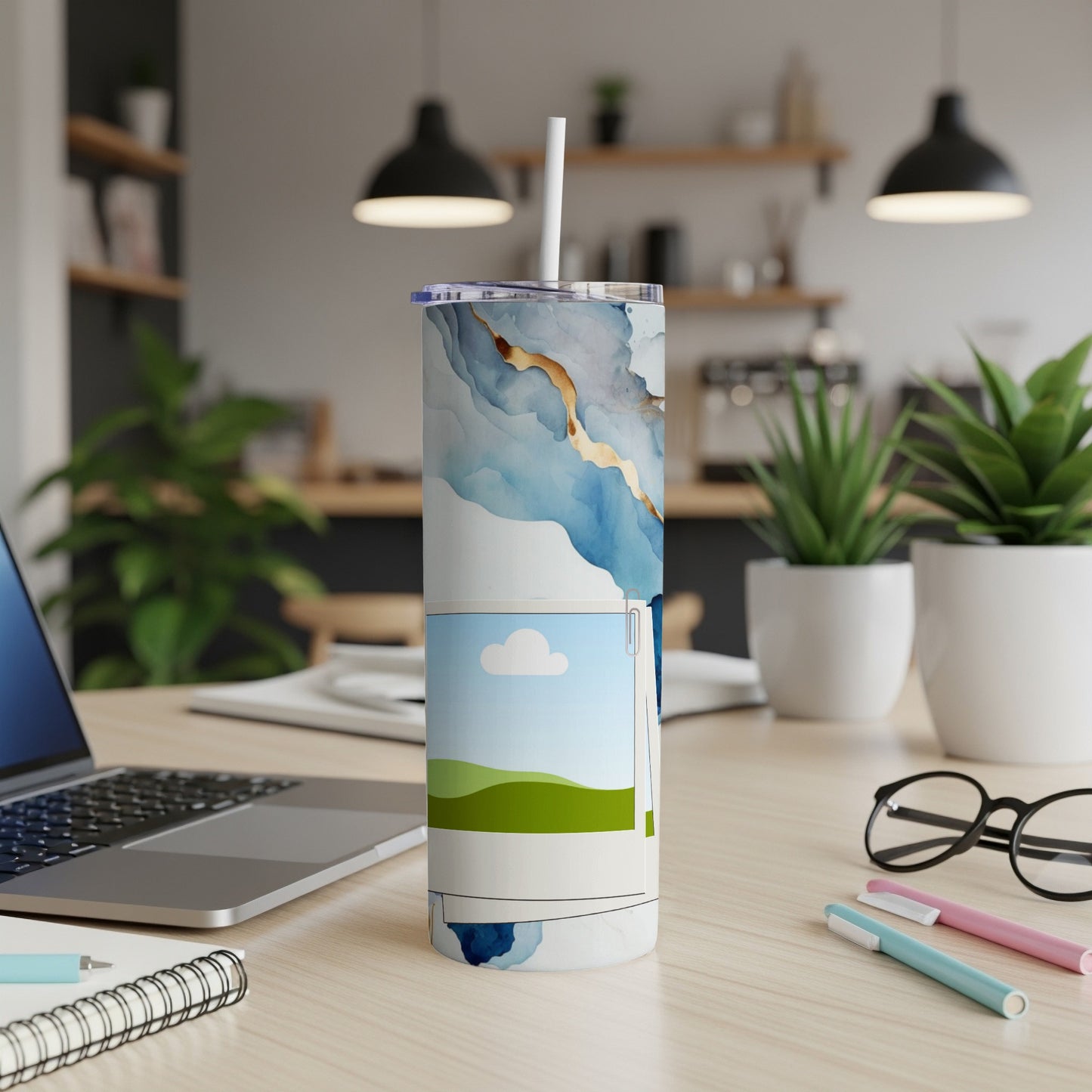 Tumbler with blue abstract art and landscape photo, straw, laptop, and pens.