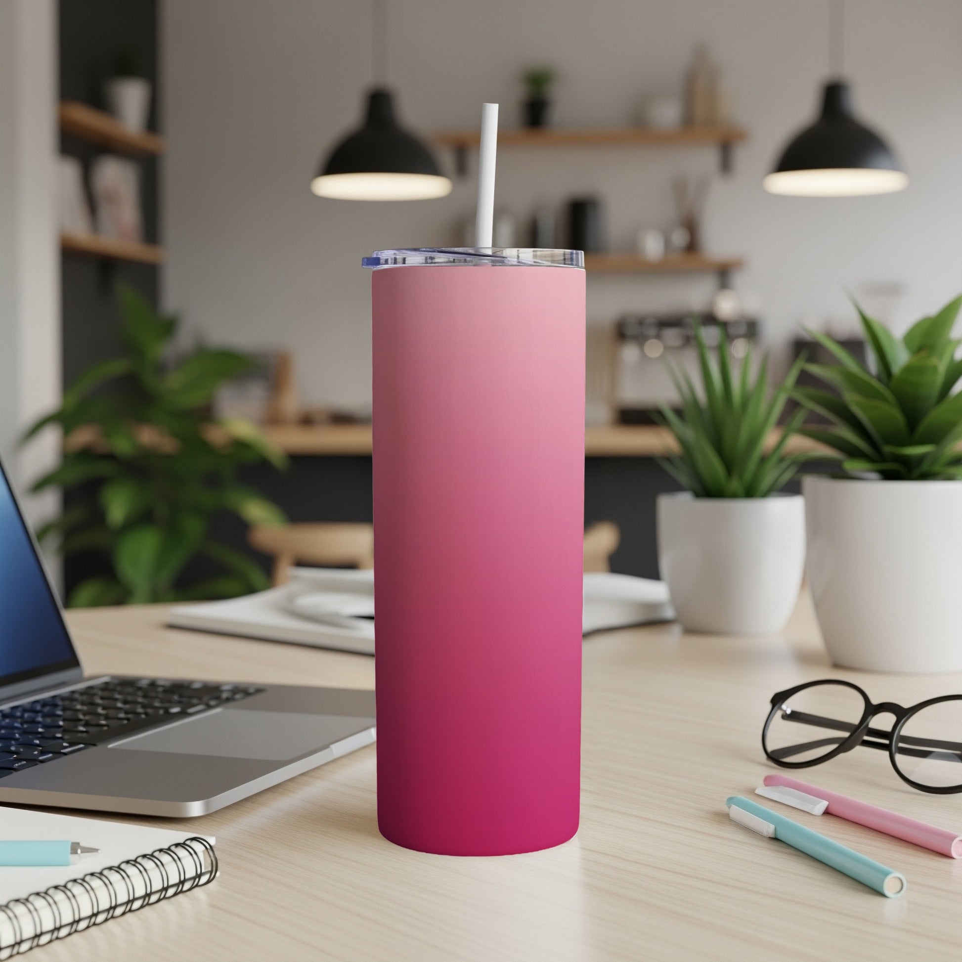Pink gradient skinny tumbler with straw on a desk next to a laptop.
