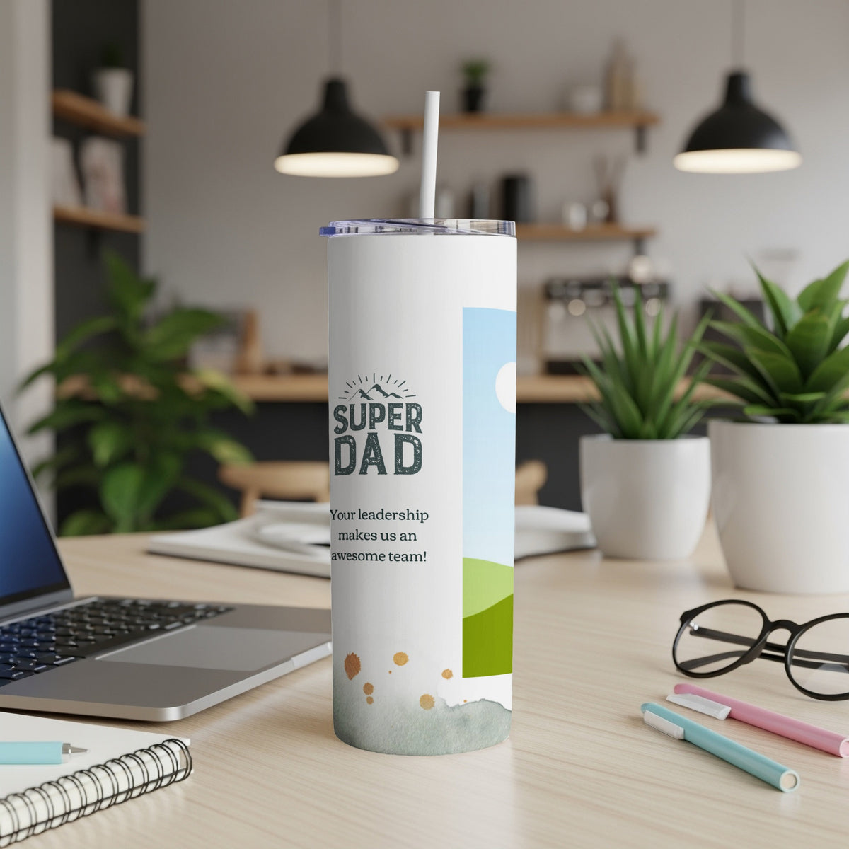 White tumbler with "Super Dad" text and landscape graphic, next to a laptop.