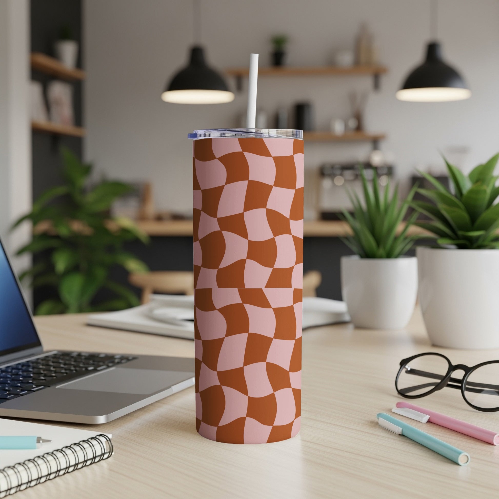 Pink and brown checkered tumbler with straw on desk.