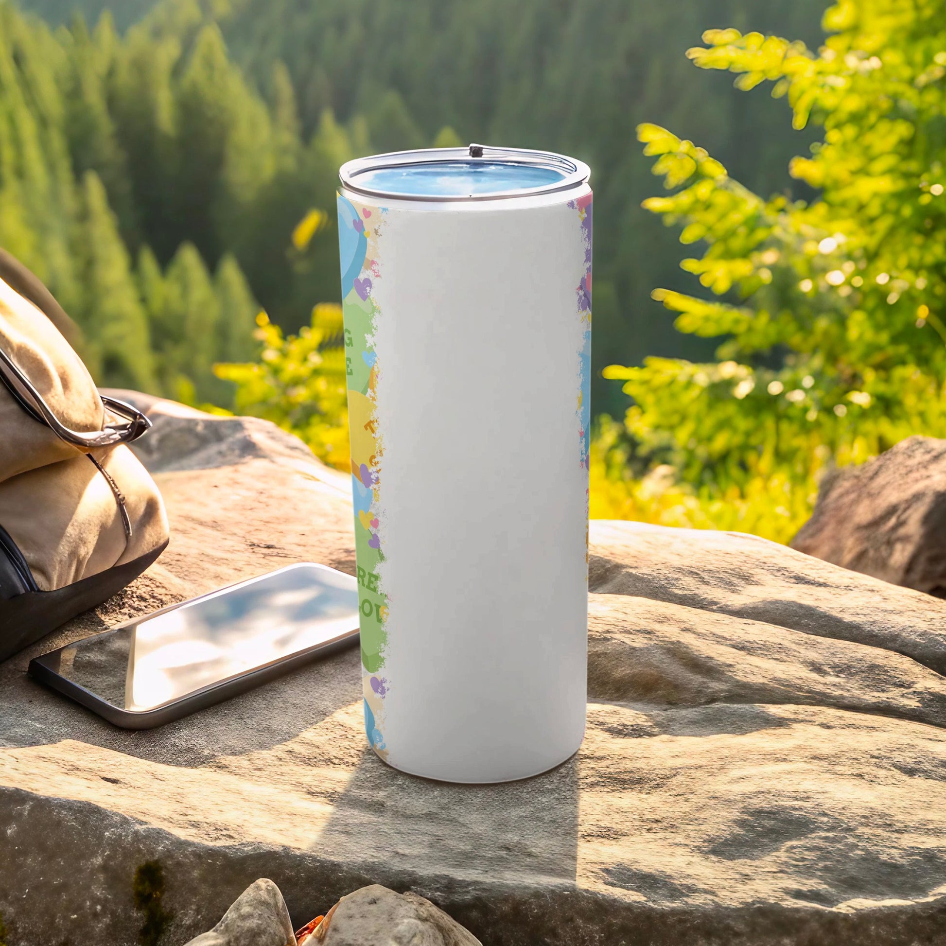 White tumbler with colorful watercolor splashes and inspirational text, on a rock with a backpack.
