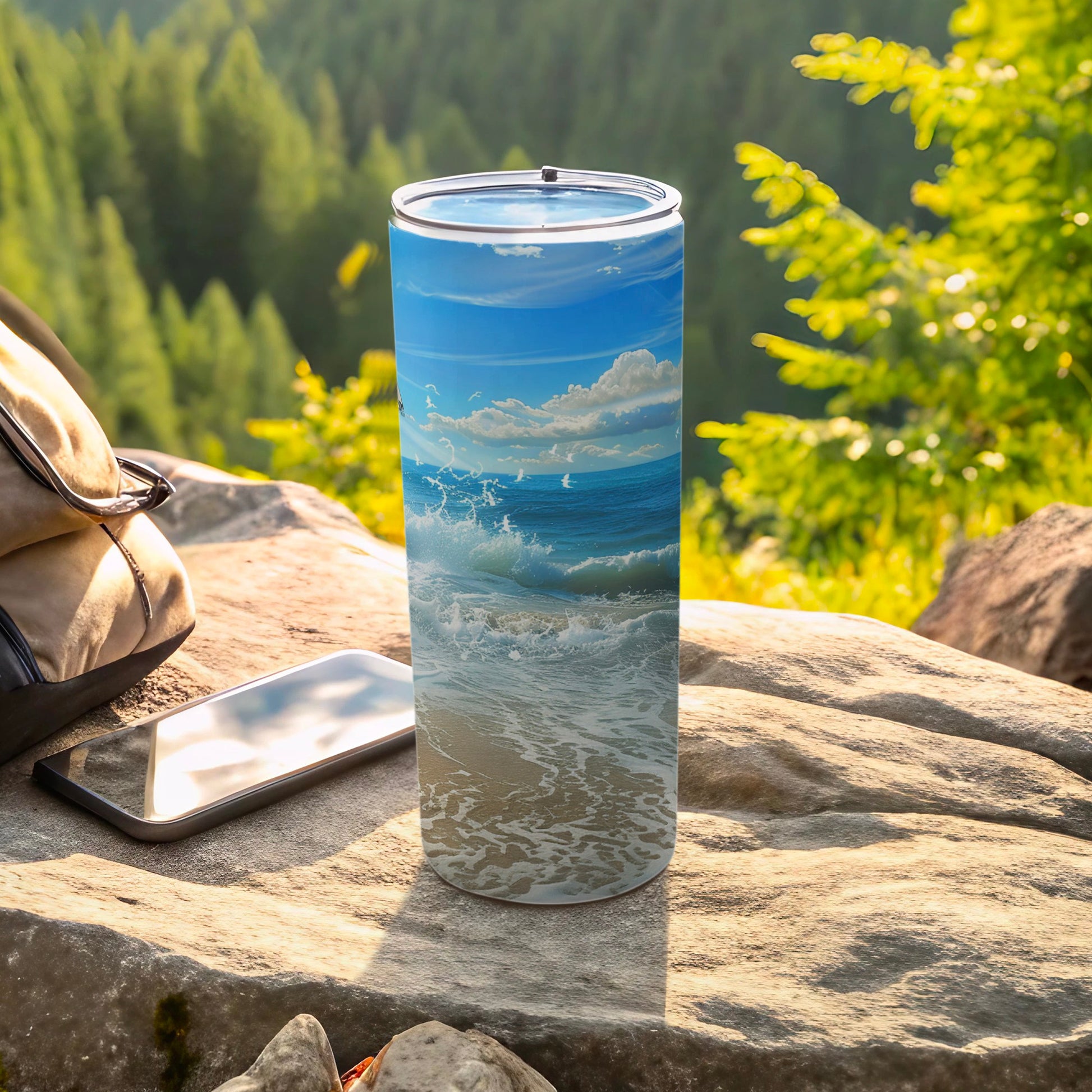 Tall tumbler with ocean wave design sits on rock next to phone and bag.