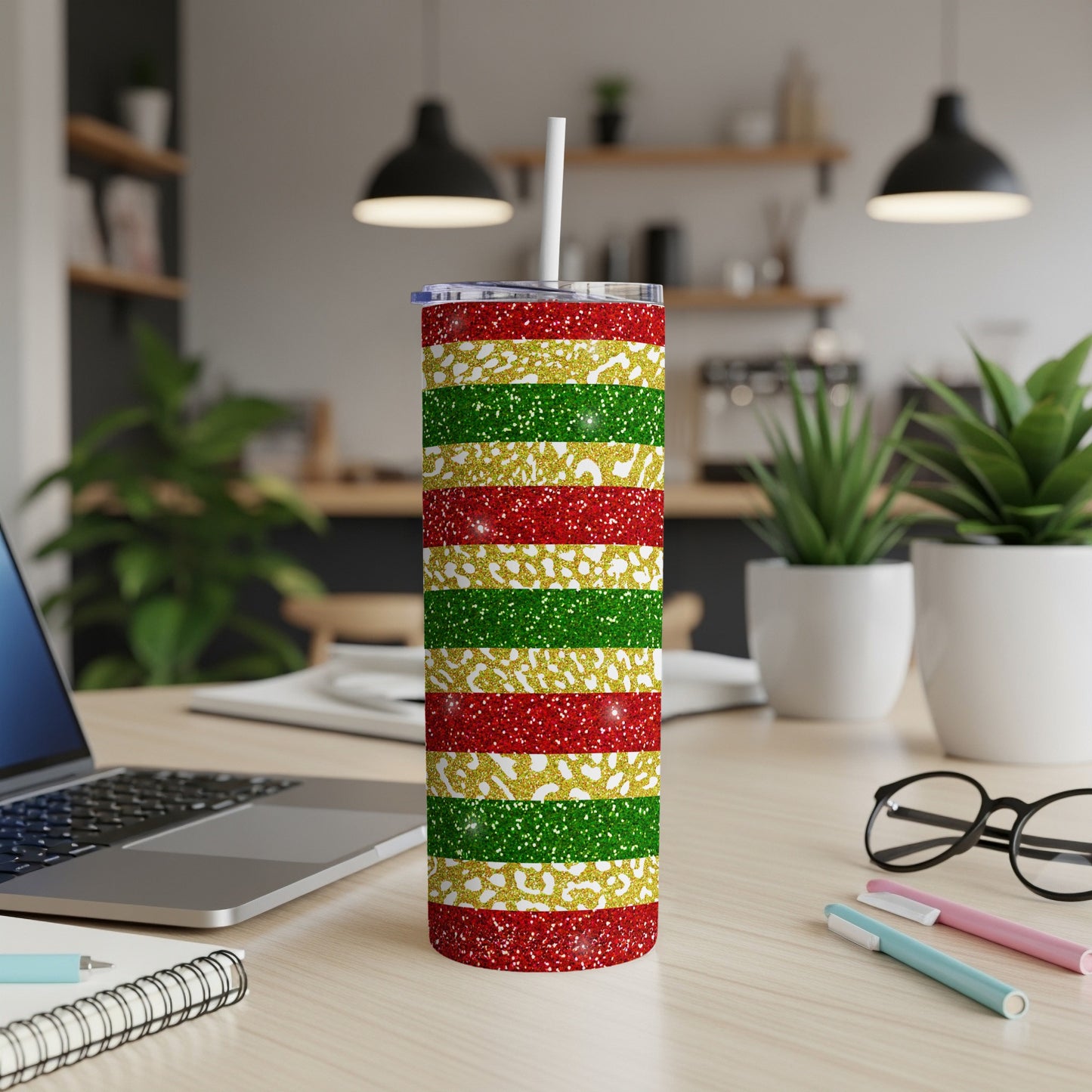 Glittering red, gold, and green striped tumbler on a desk with a laptop and pens.