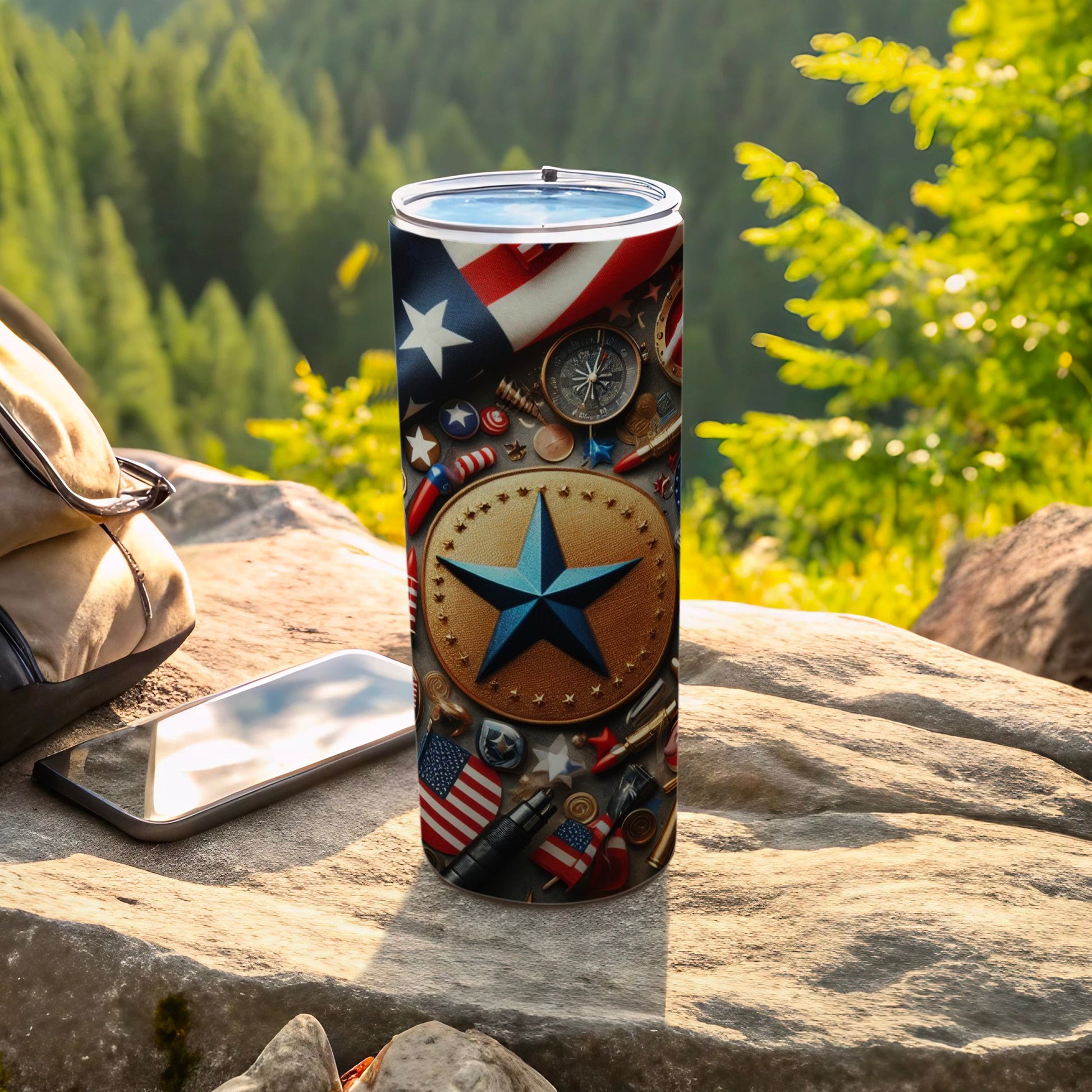 Patriotic tumbler with stars and flags, on a rock with a phone and bag.