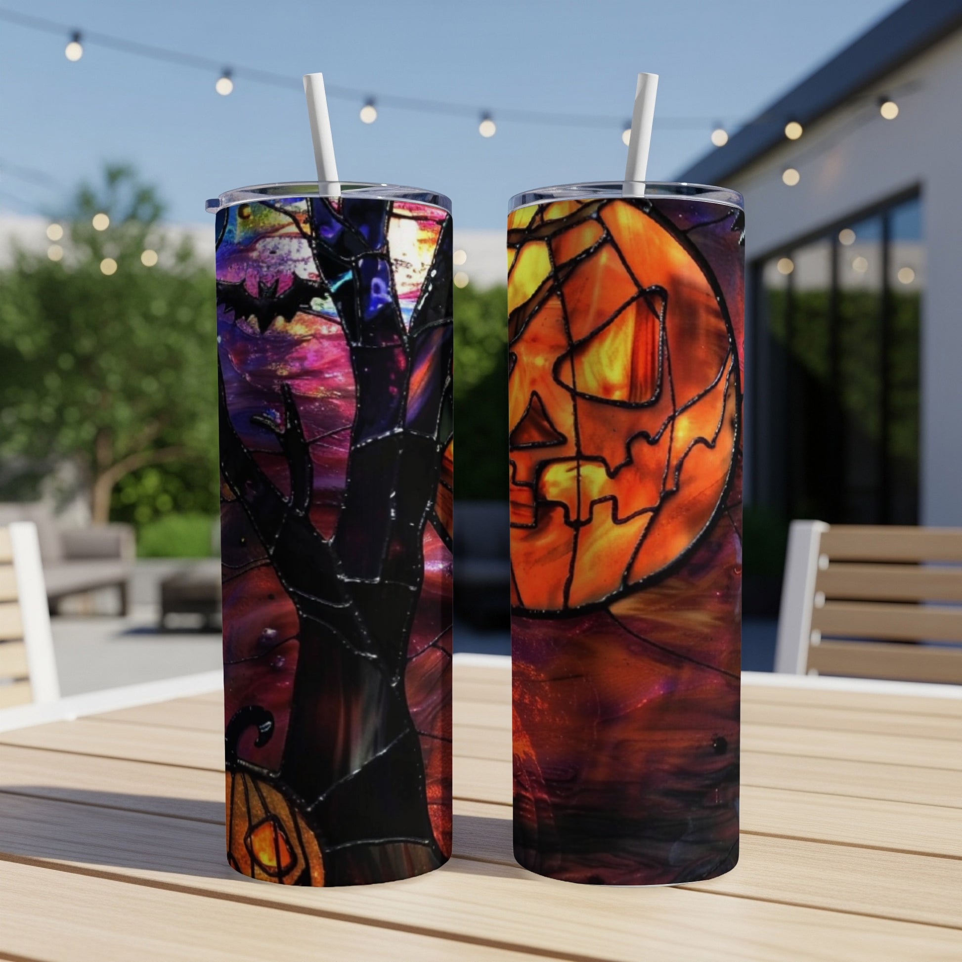 Two tumblers with Halloween jack-o'-lantern and bat designs sit on a wooden table outdoors.
