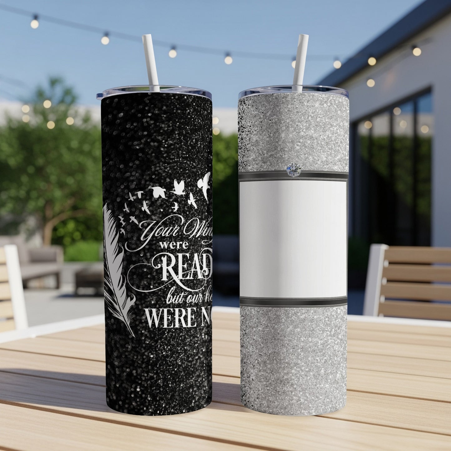Two tumblers on table: one black with white text and birds, one silver with ring design.