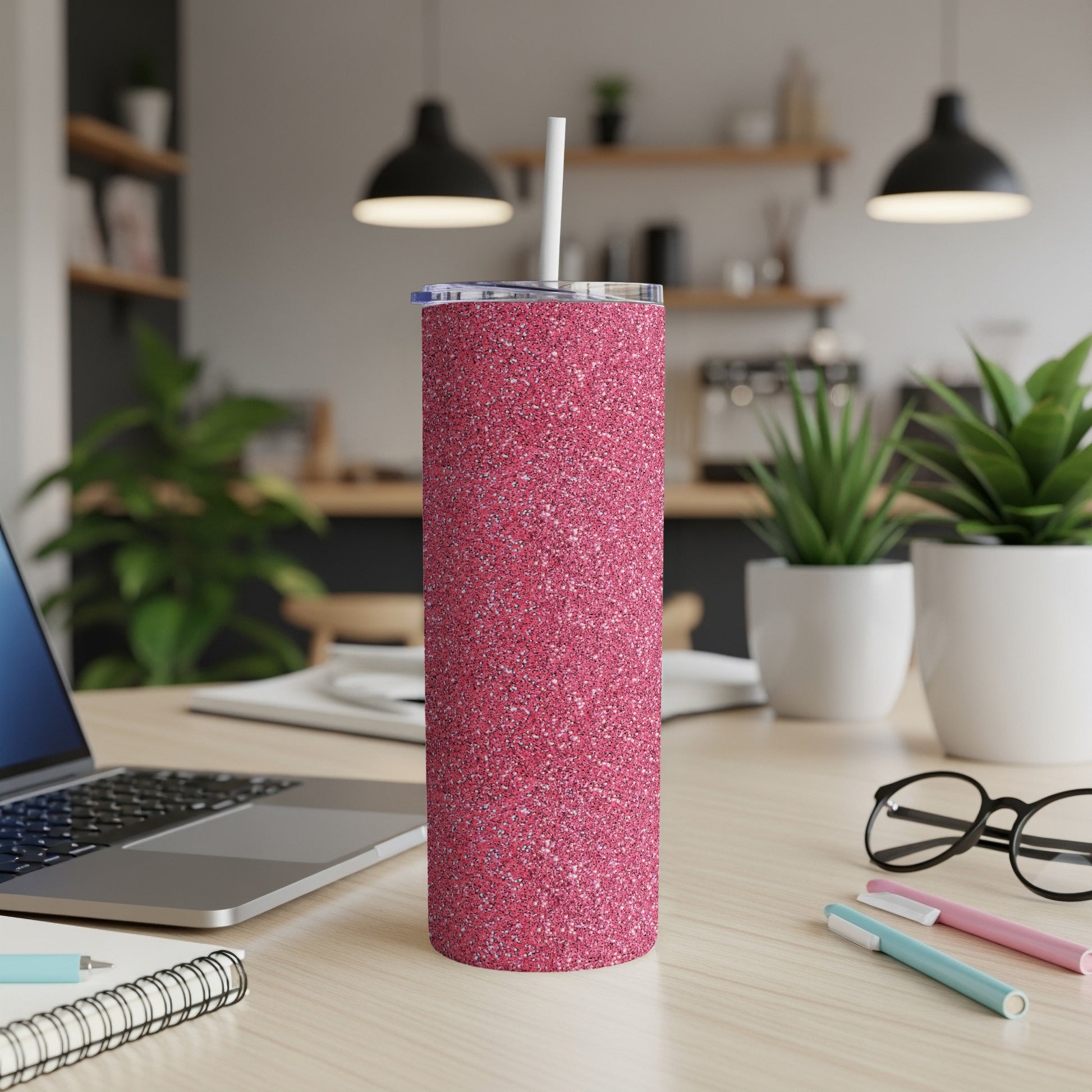 Pink glitter tumbler with straw on a desk next to laptop and notebook.