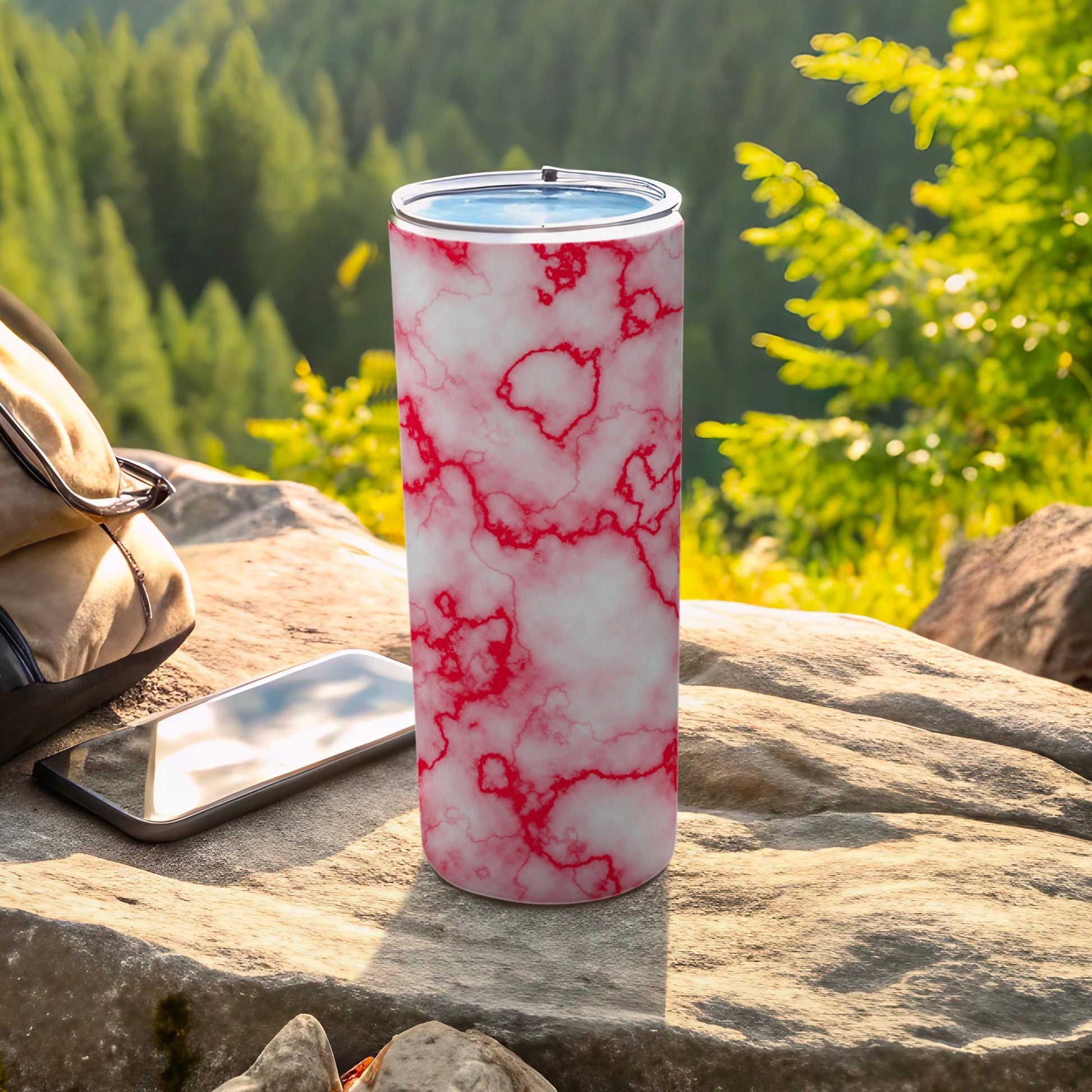 White tumbler with red marble pattern on a rock with phone and backpack.