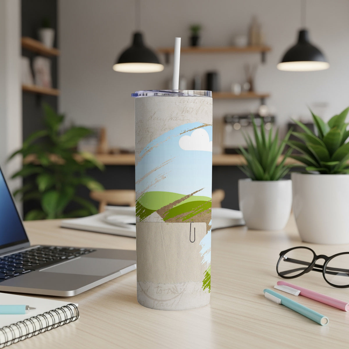 Tall tumbler with a landscape design, straw, and paperclip detail on a desk.