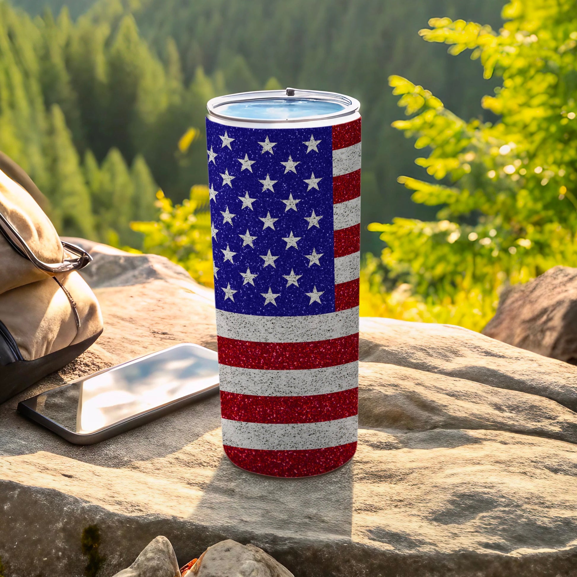 Glittery American flag tumbler on rock with phone and backpack.