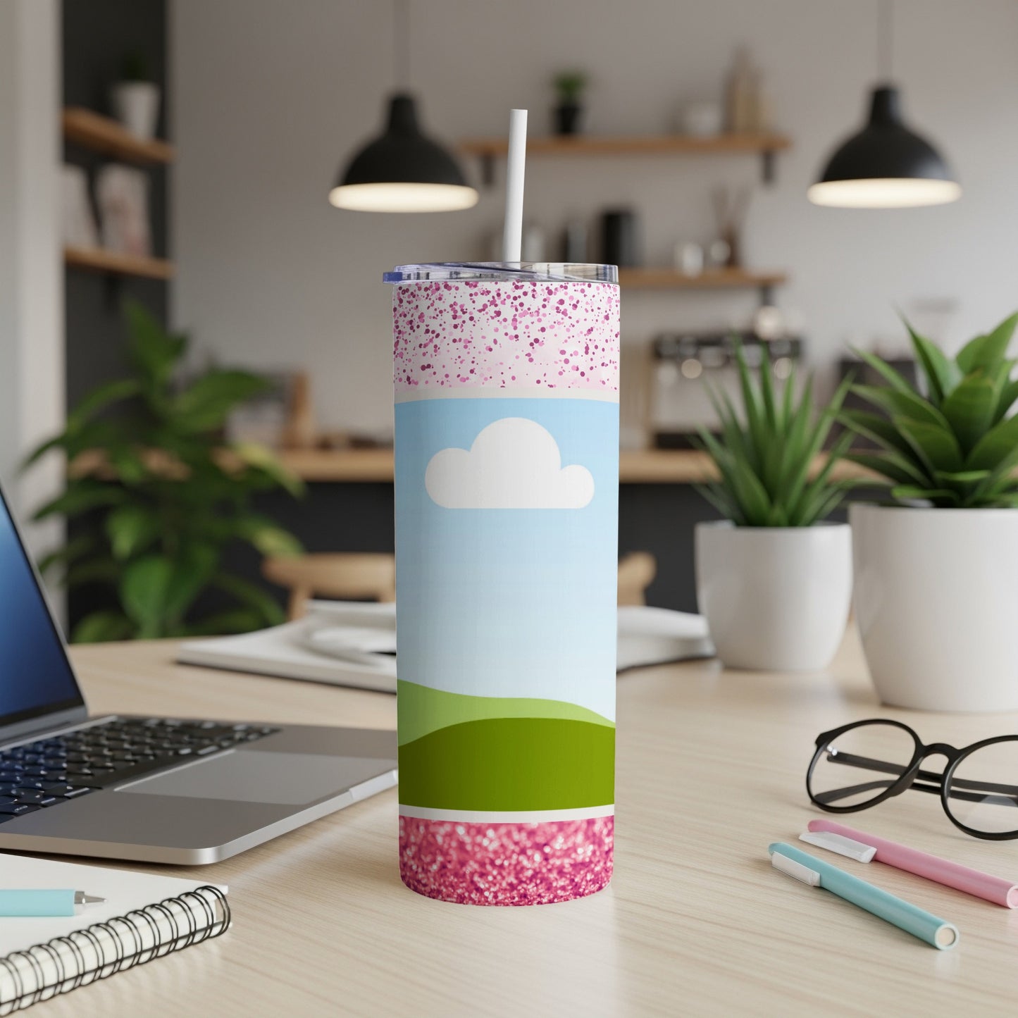 Tumbler with pink glitter, sky, and green hills design on a desk.