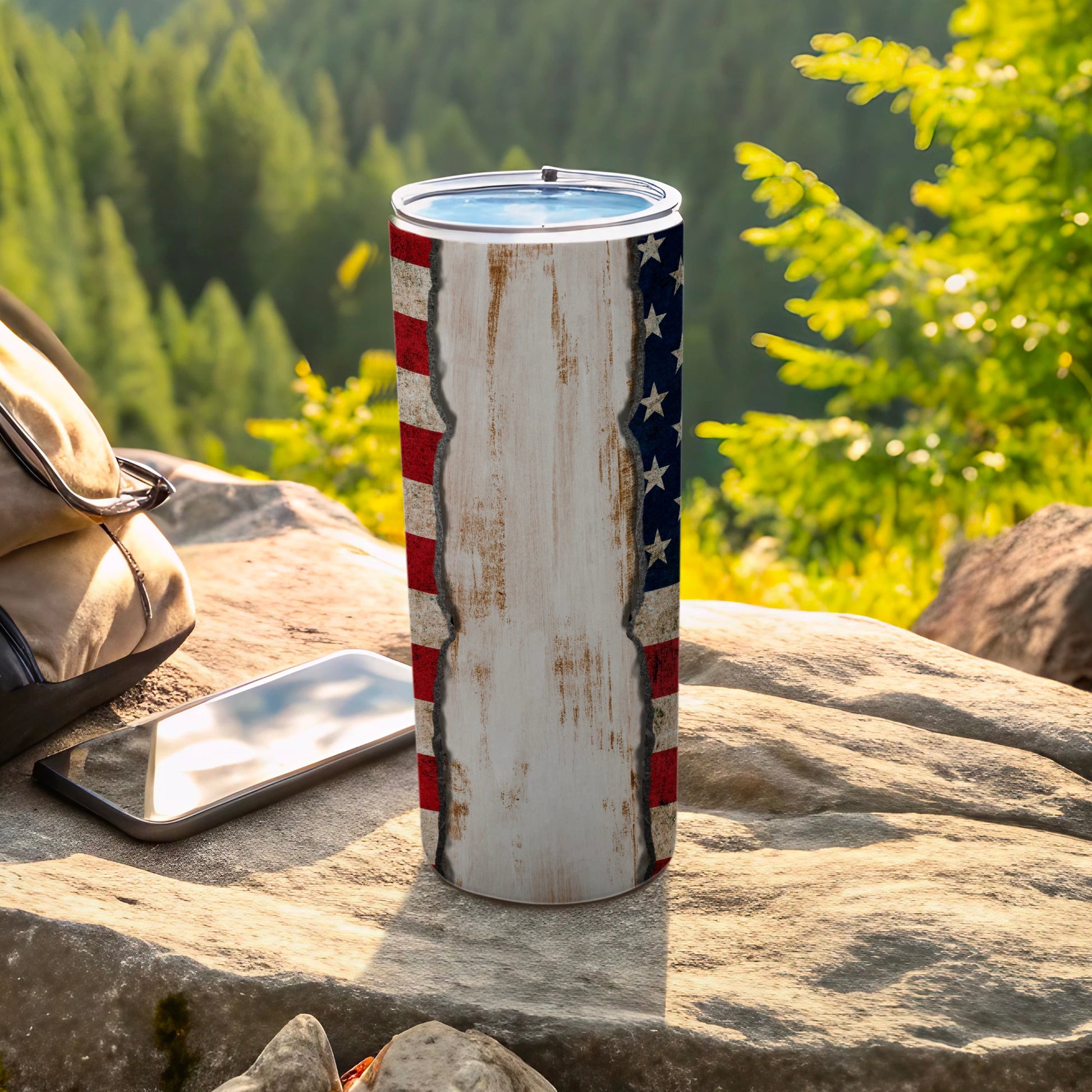 Tumbler with American flag design, rests on rocks near a backpack and phone.