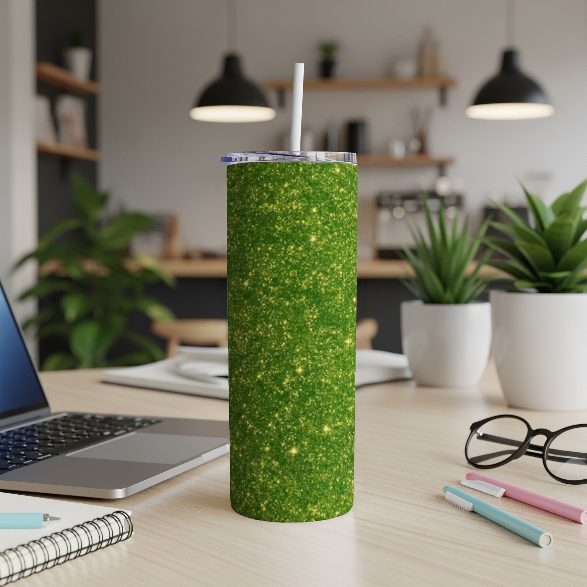 Green glitter tumbler with straw on a desk with laptop and plants.