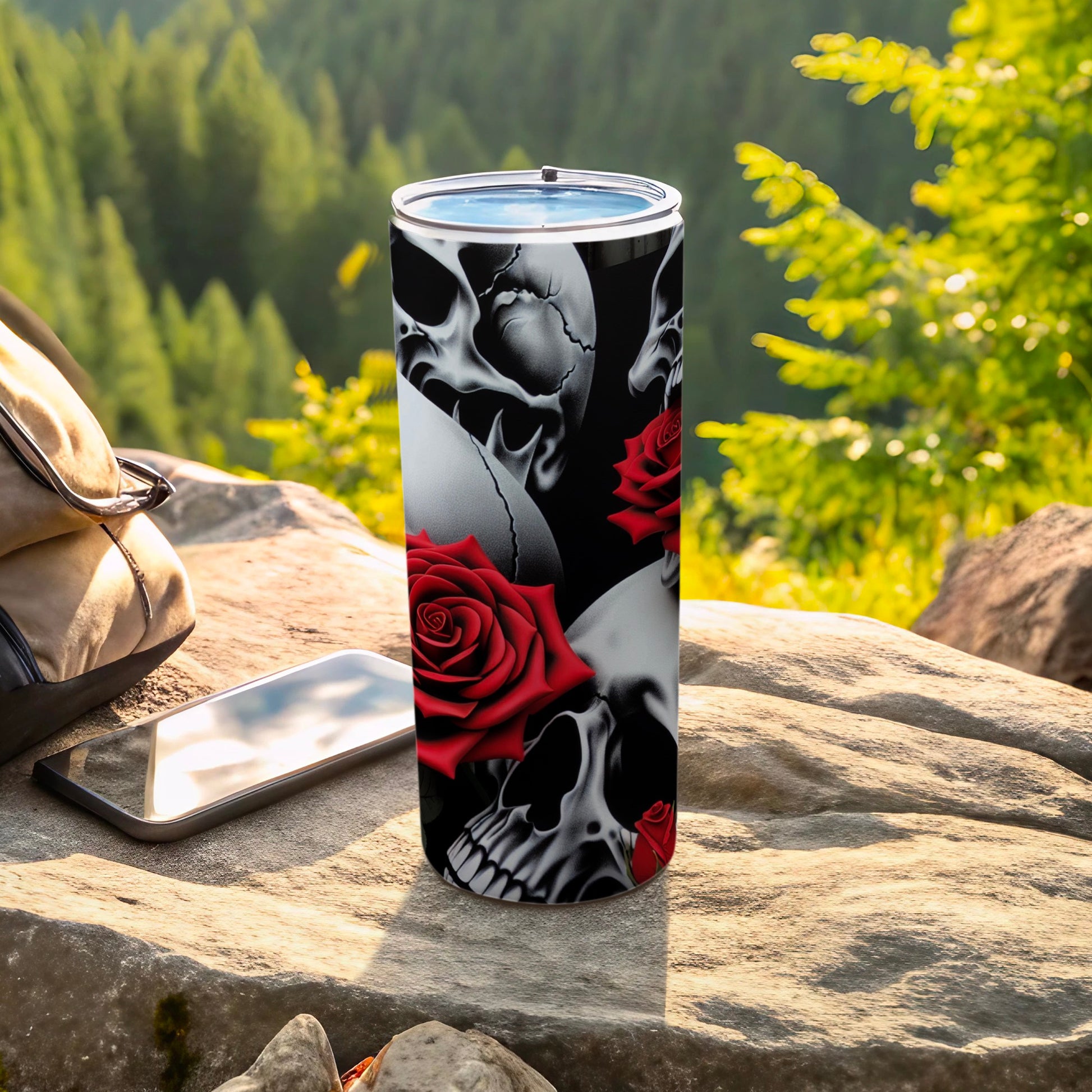 Skulls and red roses adorn a stainless steel tumbler on a rocky surface outdoors.