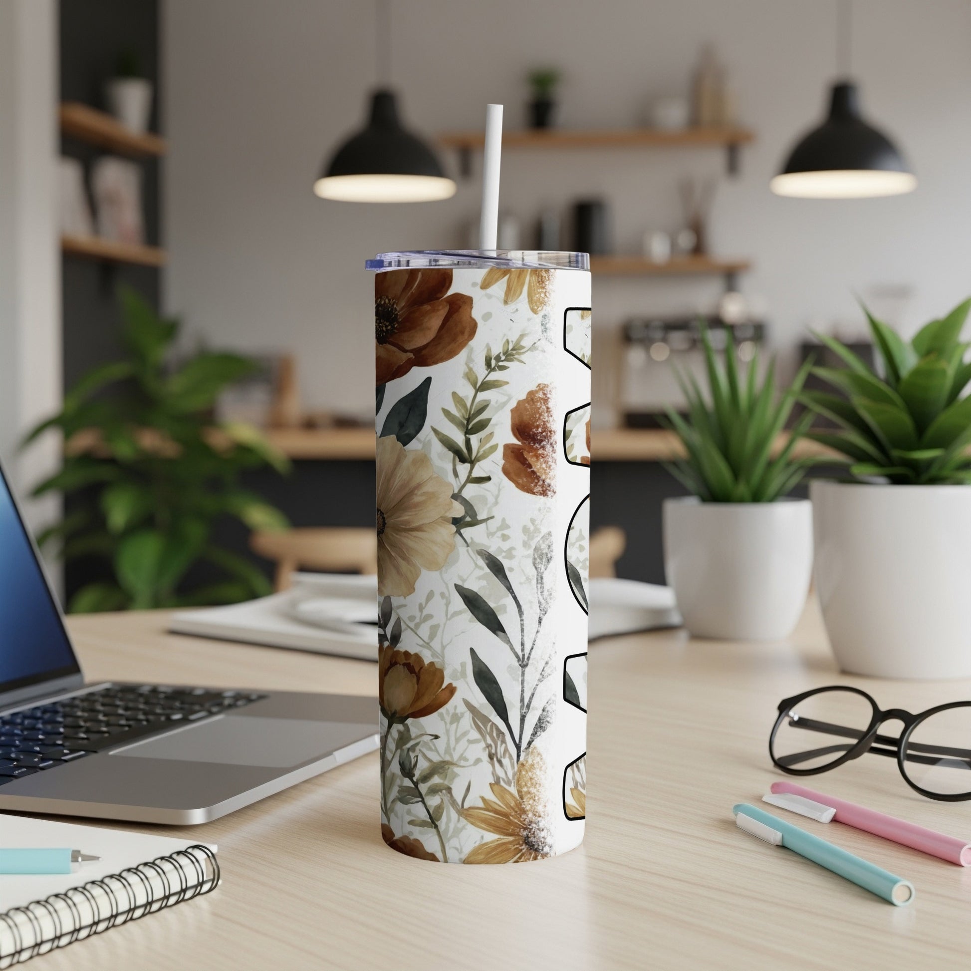 Floral patterned tumbler with straw on desk next to laptop and pens.